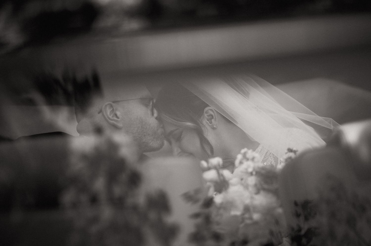 Black and white photo of bride and groom sharing a kiss inside a car on their wedding day.