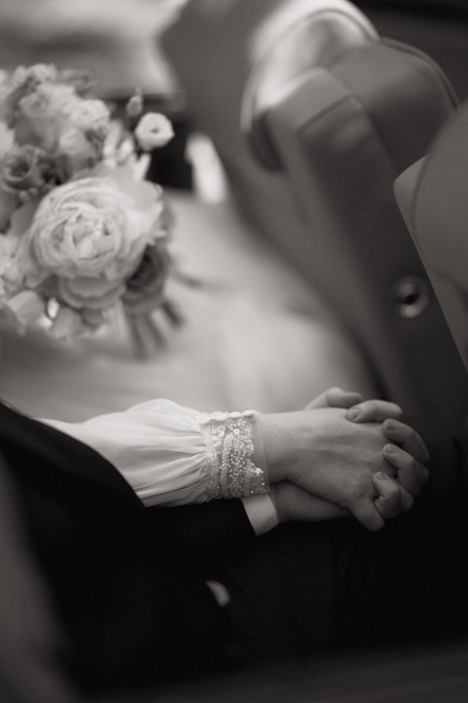 Black and white photo of bride and groom holding hands, featuring lace bridal sleeve and wedding bouquet.