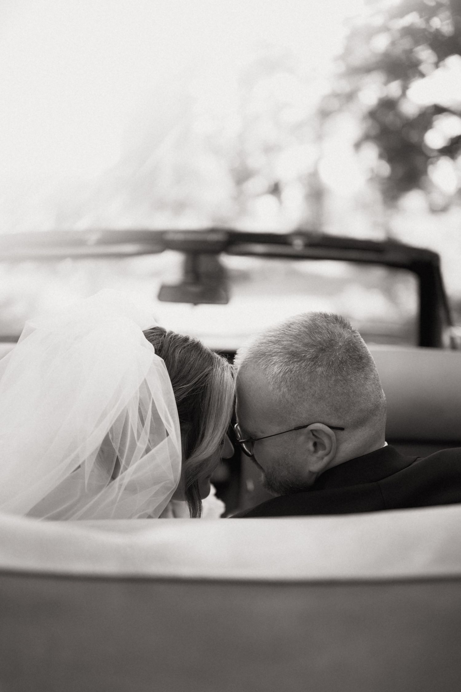 Black and white photo of bride and groom sharing a kiss in a convertible car on their wedding day.