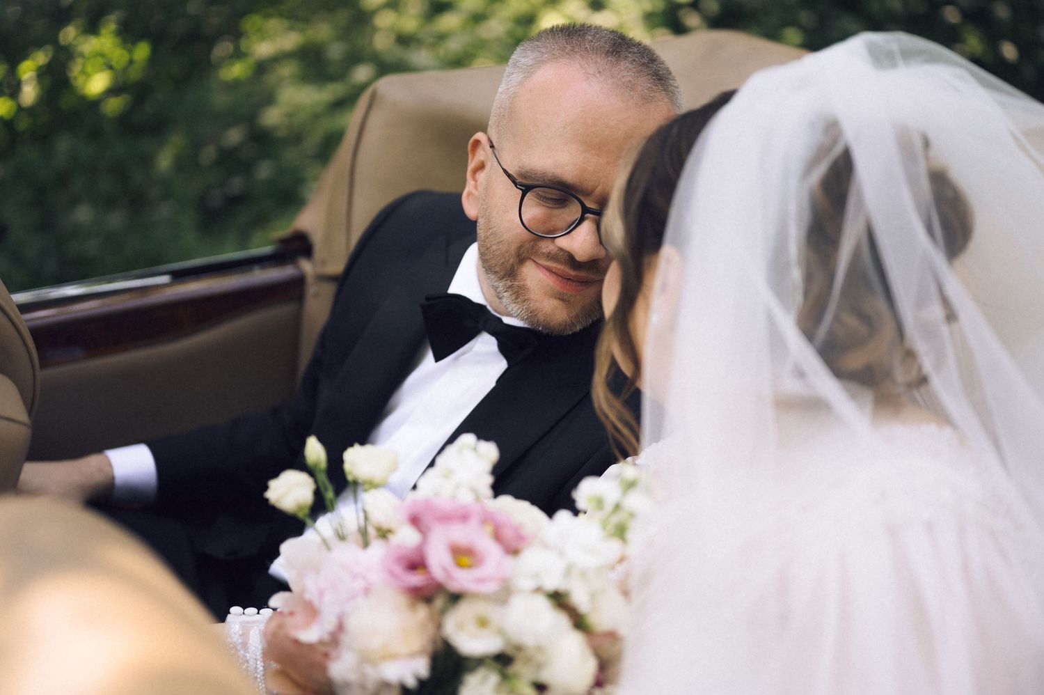Groom in tuxedo and bow tie smiling at bride holding pink and white wedding bouquet in vintage car.