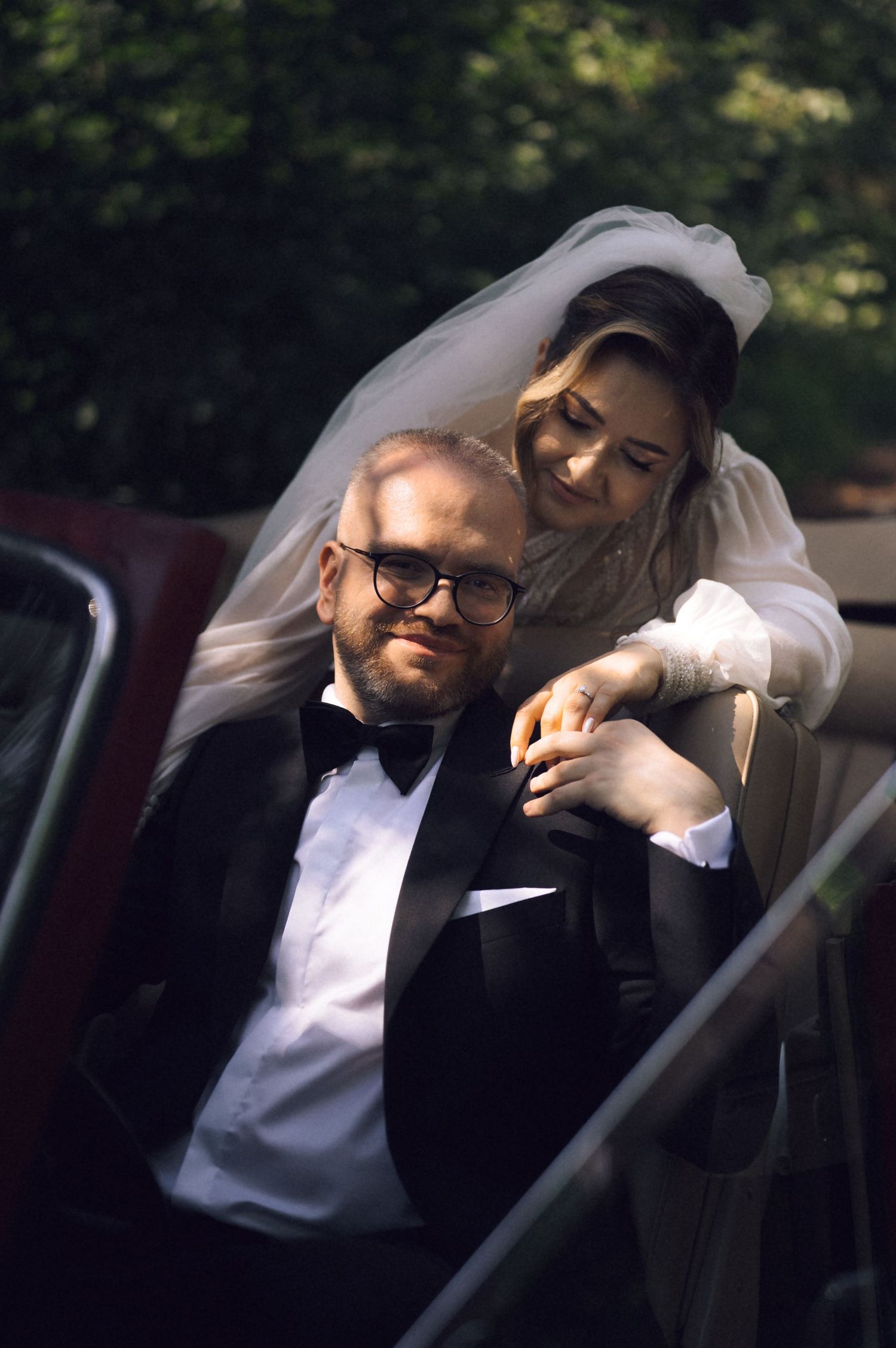 A groom in a tuxedo sits by a vintage car while his bride in a veil leans over him tenderly on their wedding day.