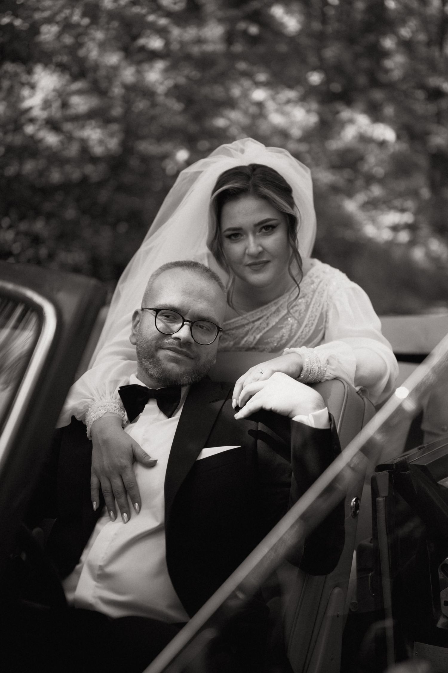 Black and white wedding photo of bride in veil embracing groom in tuxedo and bow tie beside a vintage car.