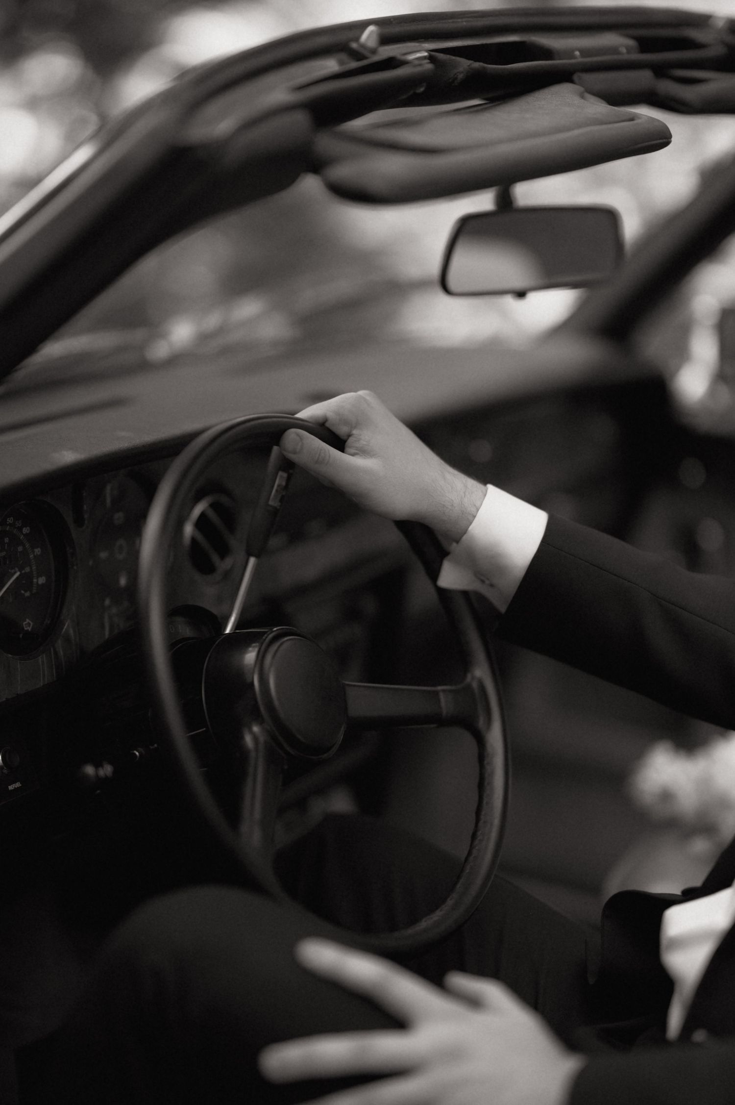 Groom in black suit gripping vintage car steering wheel, black and white wedding photo.