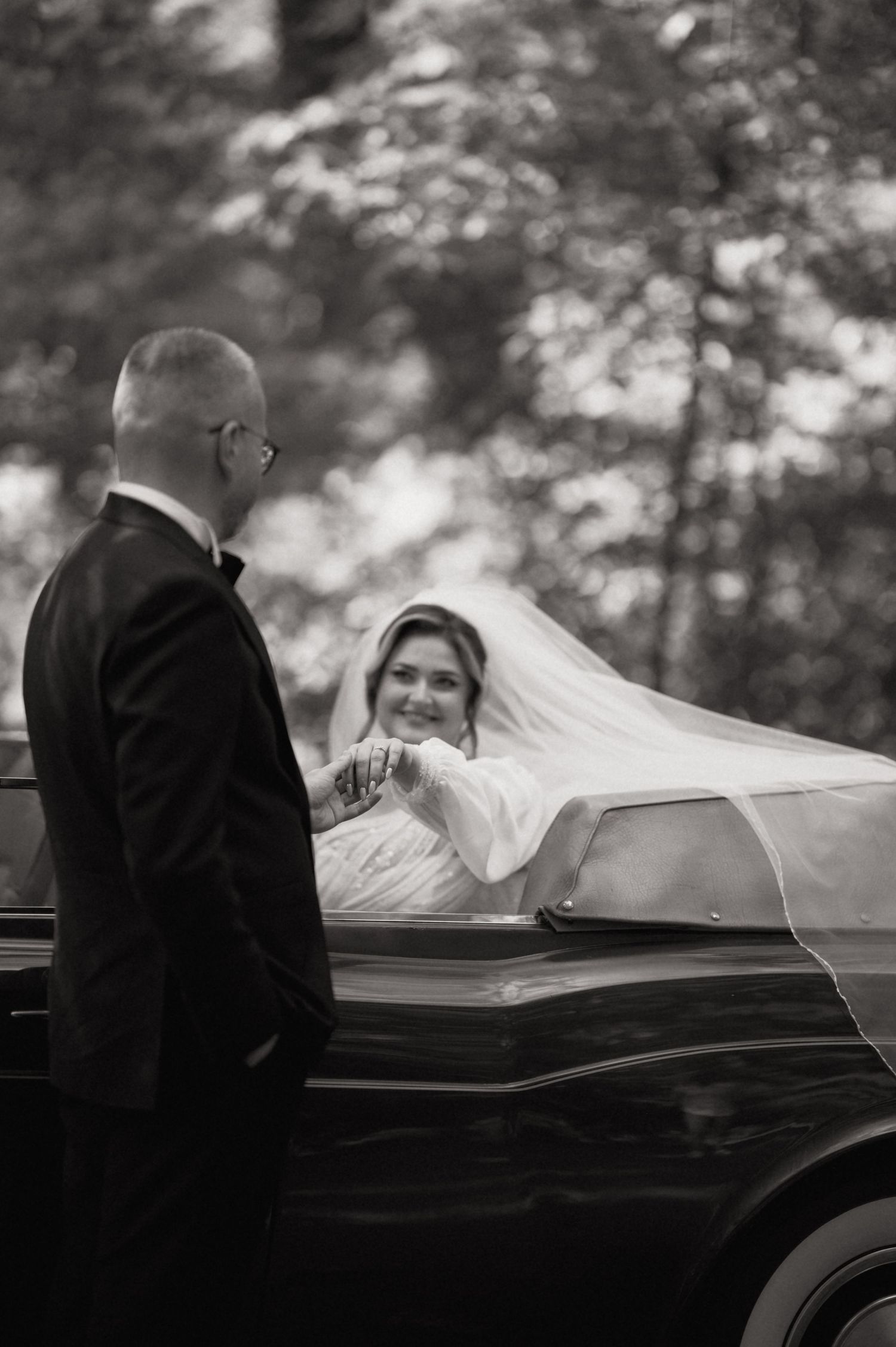 Smiling bride in white gown and veil seated in classic car, greeting groom outside.