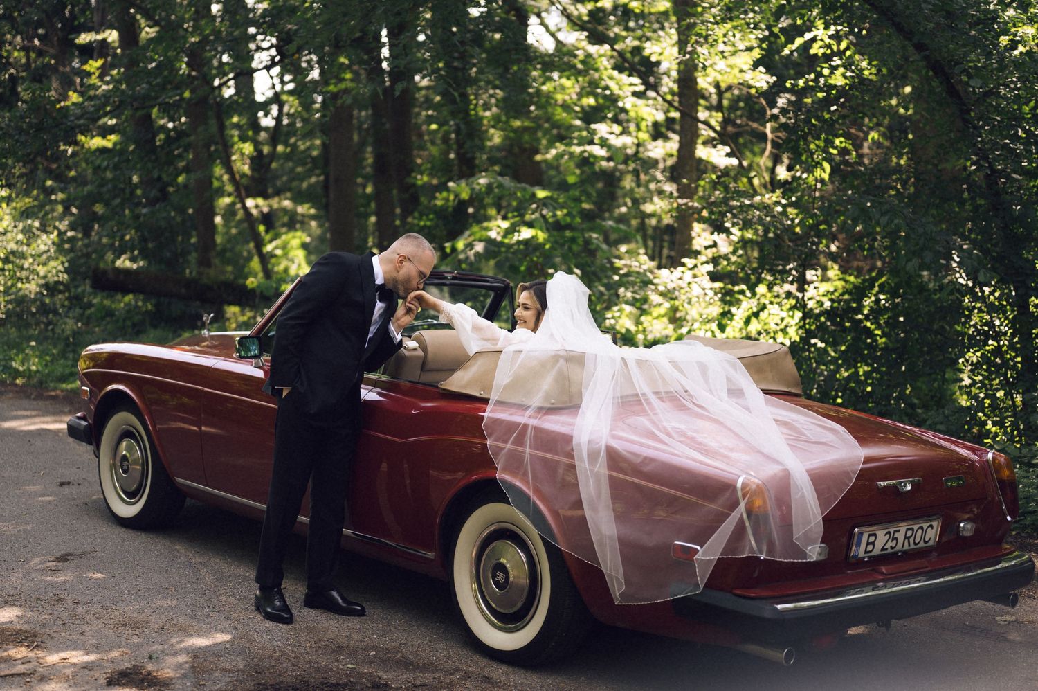 Bride in white gown and veil sits in vintage red convertible as groom in tuxedo leans in for a kiss.