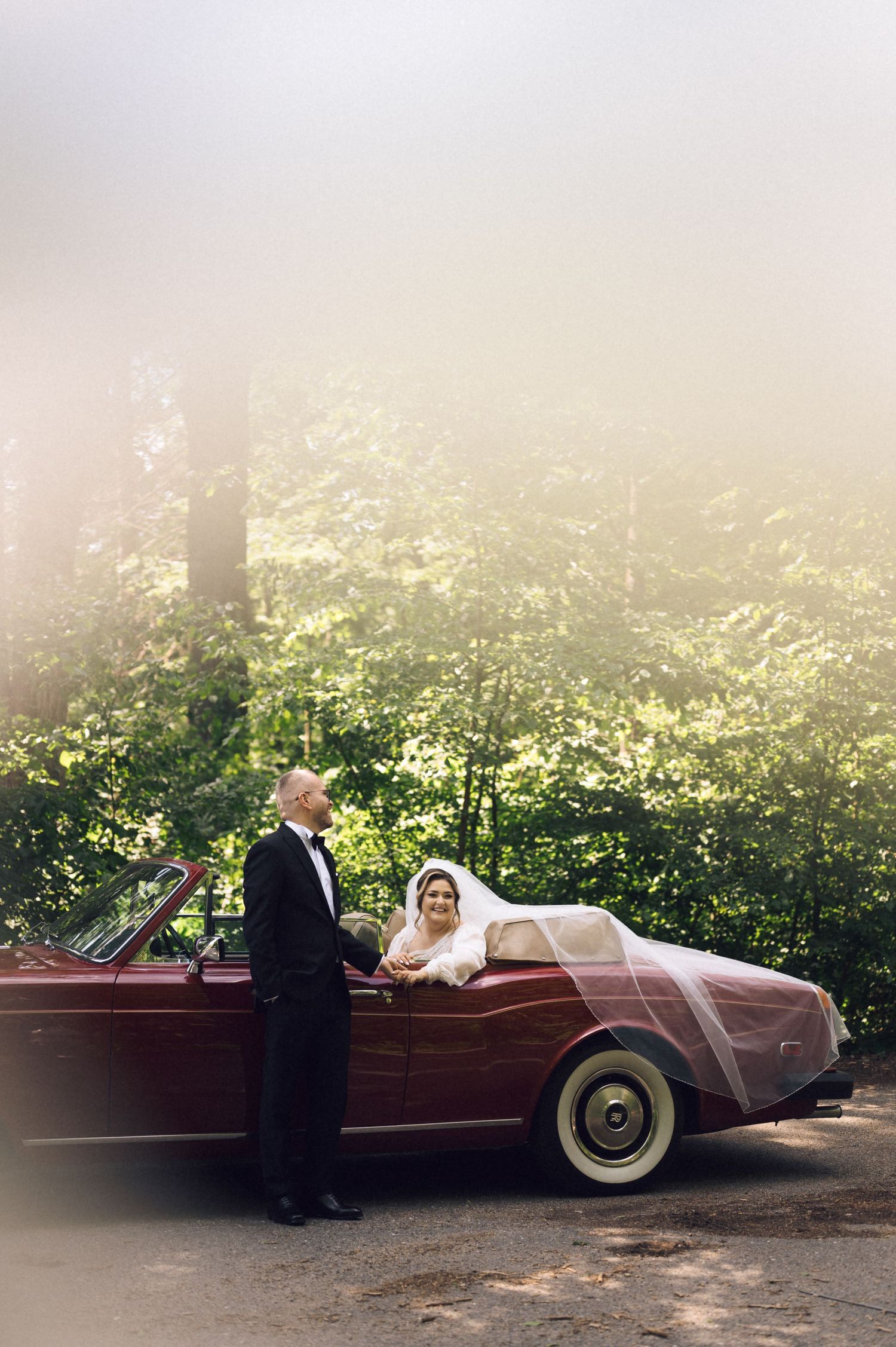Bride in white gown and veil leans on vintage red convertible as groom in tuxedo stands beside her in misty forest.
