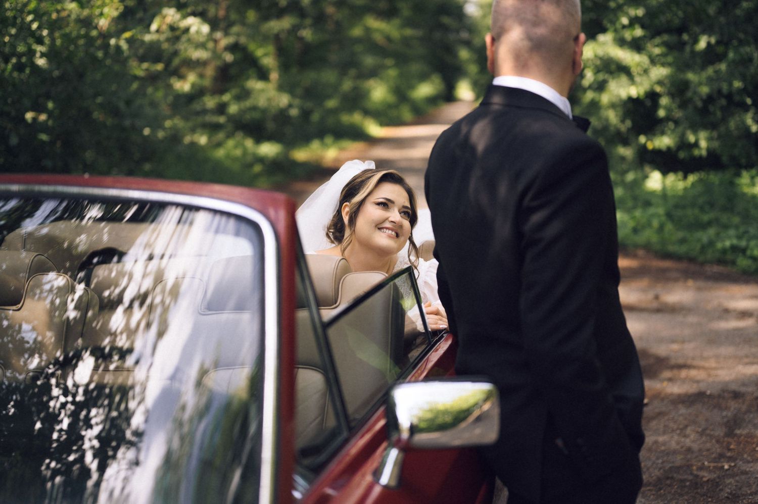 Bride smiling at groom beside a vintage red car on a tree-lined path on their wedding day.