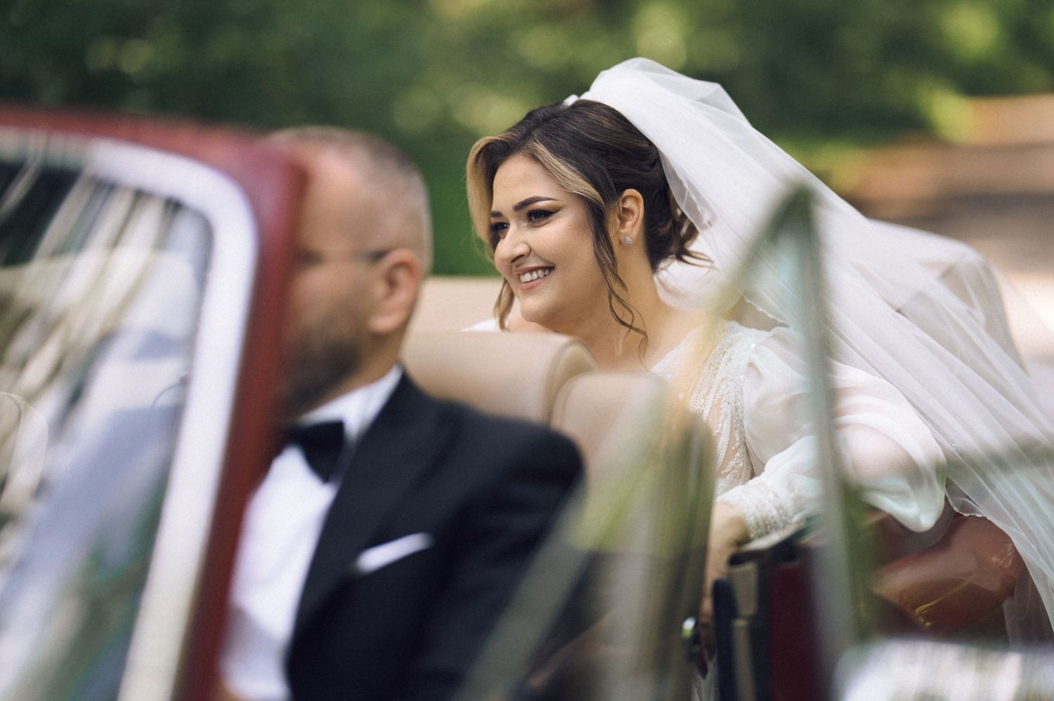 Smiling bride in white veil sits in vintage red car beside groom in black tuxedo on wedding day.