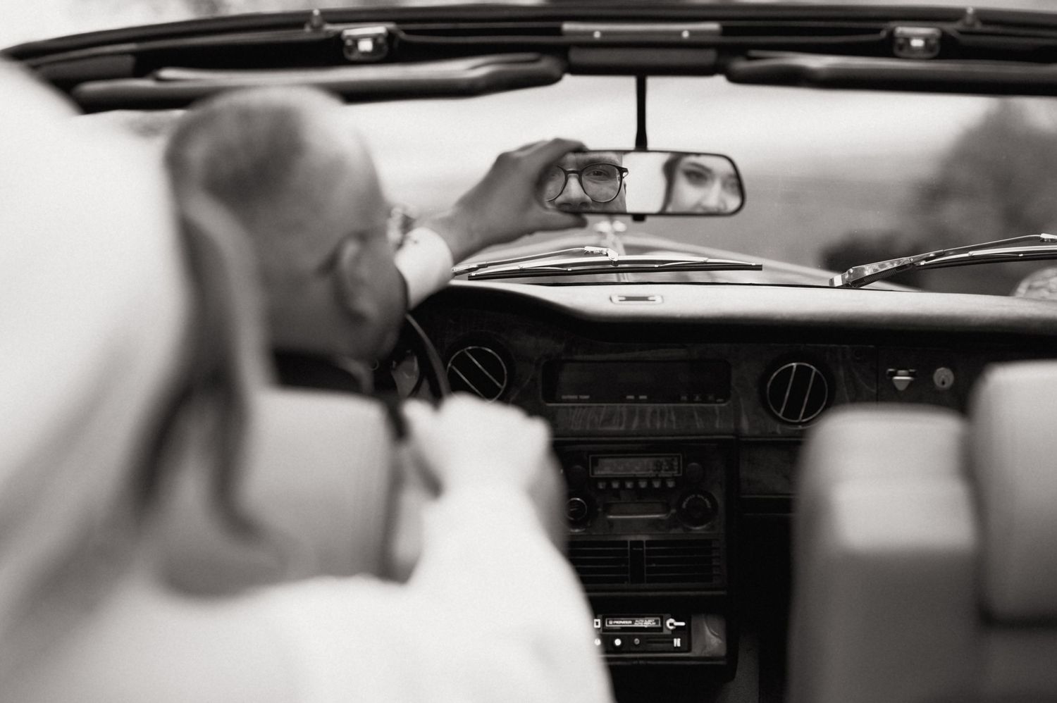 Black and white photo of a man adjusting a rearview mirror inside a classic convertible car.
