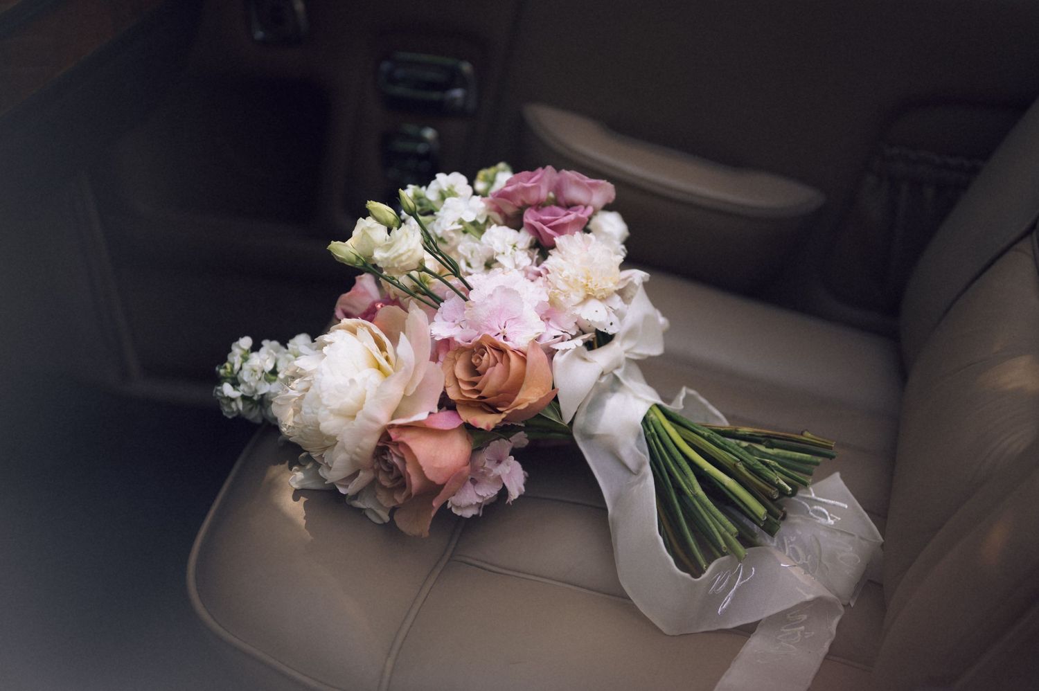 A bridal bouquet of pink, white, and peach roses resting on a beige leather car seat.