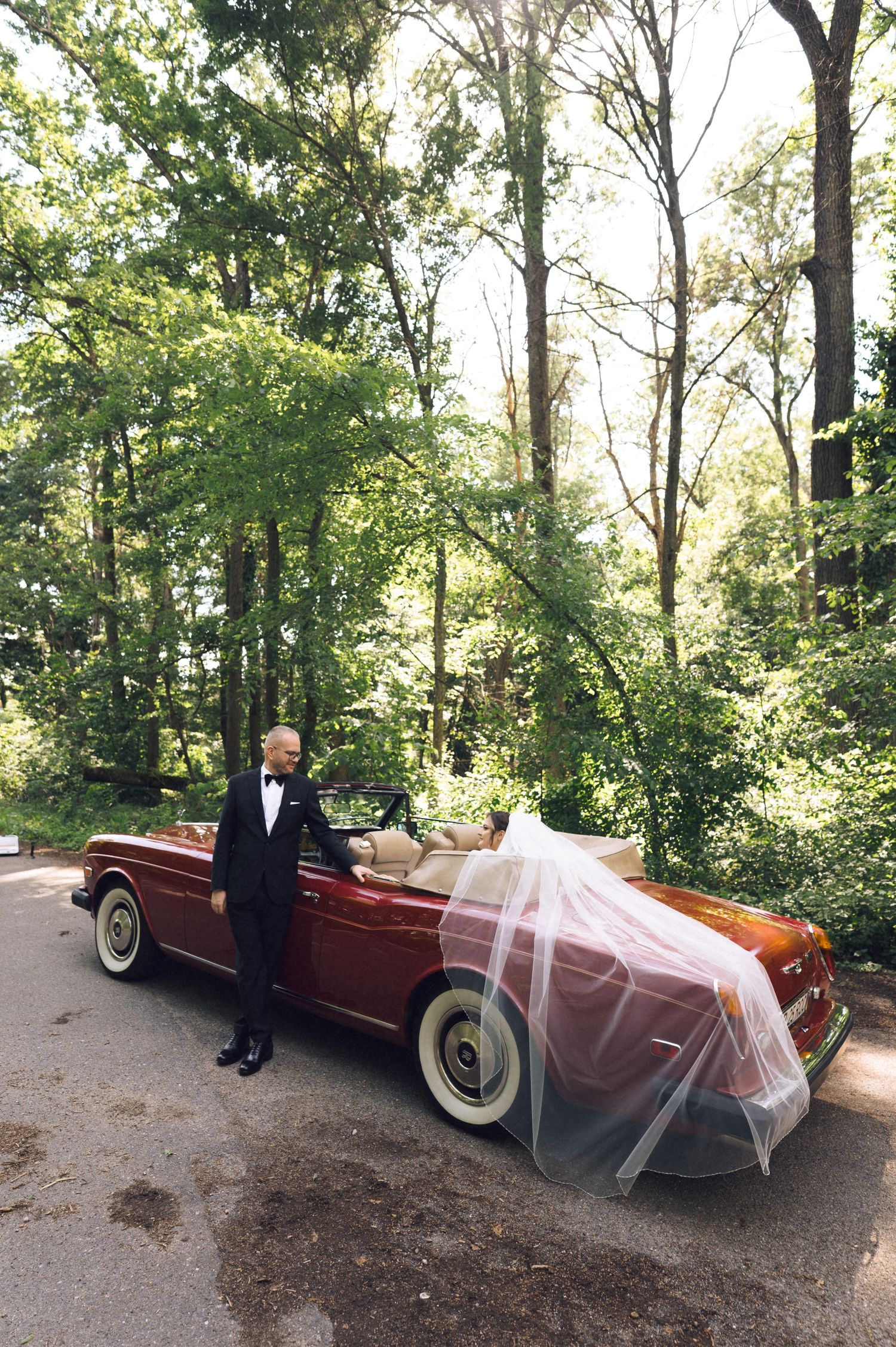 Bride and groom posing with a vintage red convertible car surrounded by lush green trees on their wedding day.