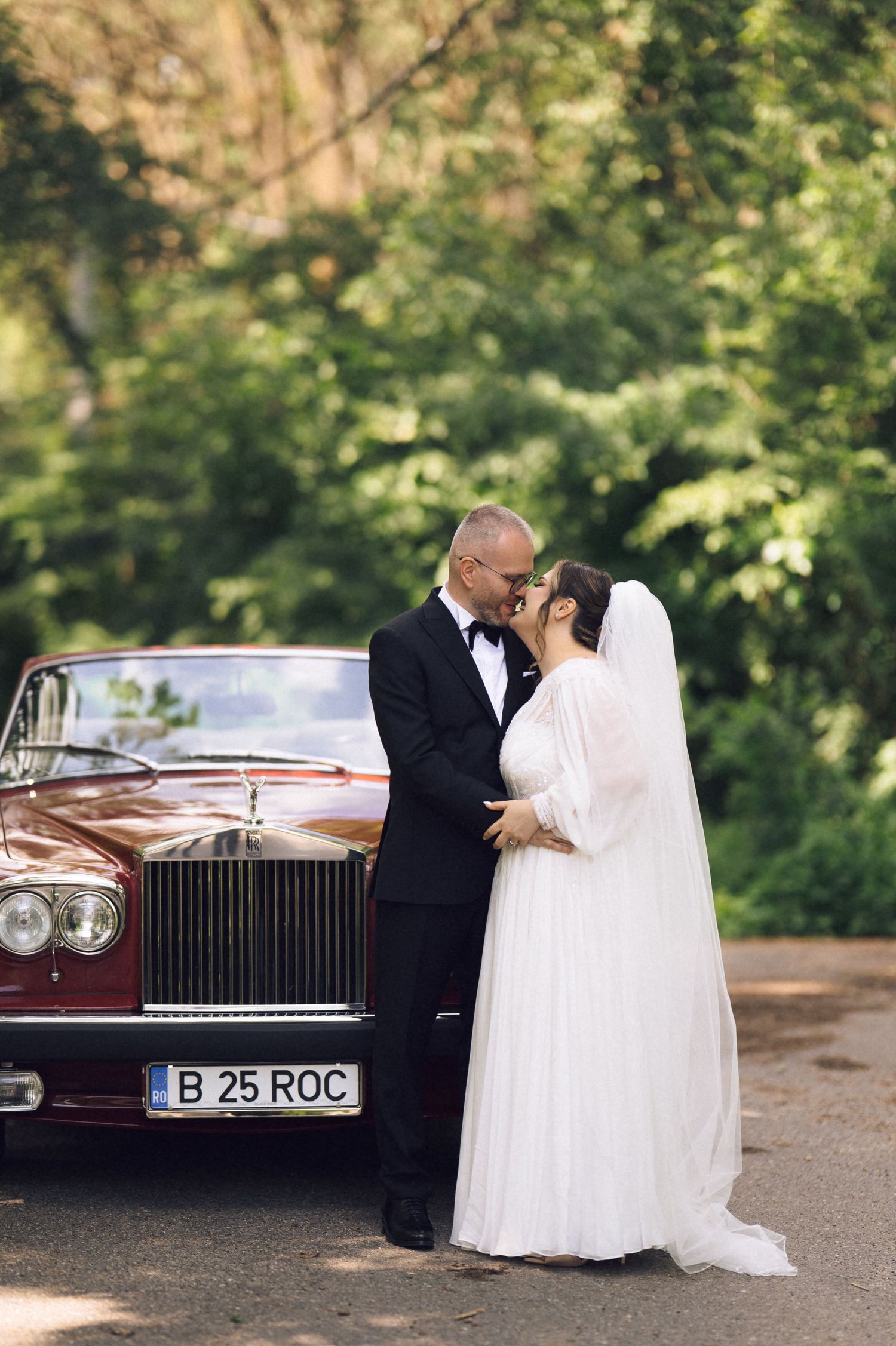 Bride and groom kissing beside a vintage red Rolls-Royce with license plate B25ROC, surrounded by lush green trees.