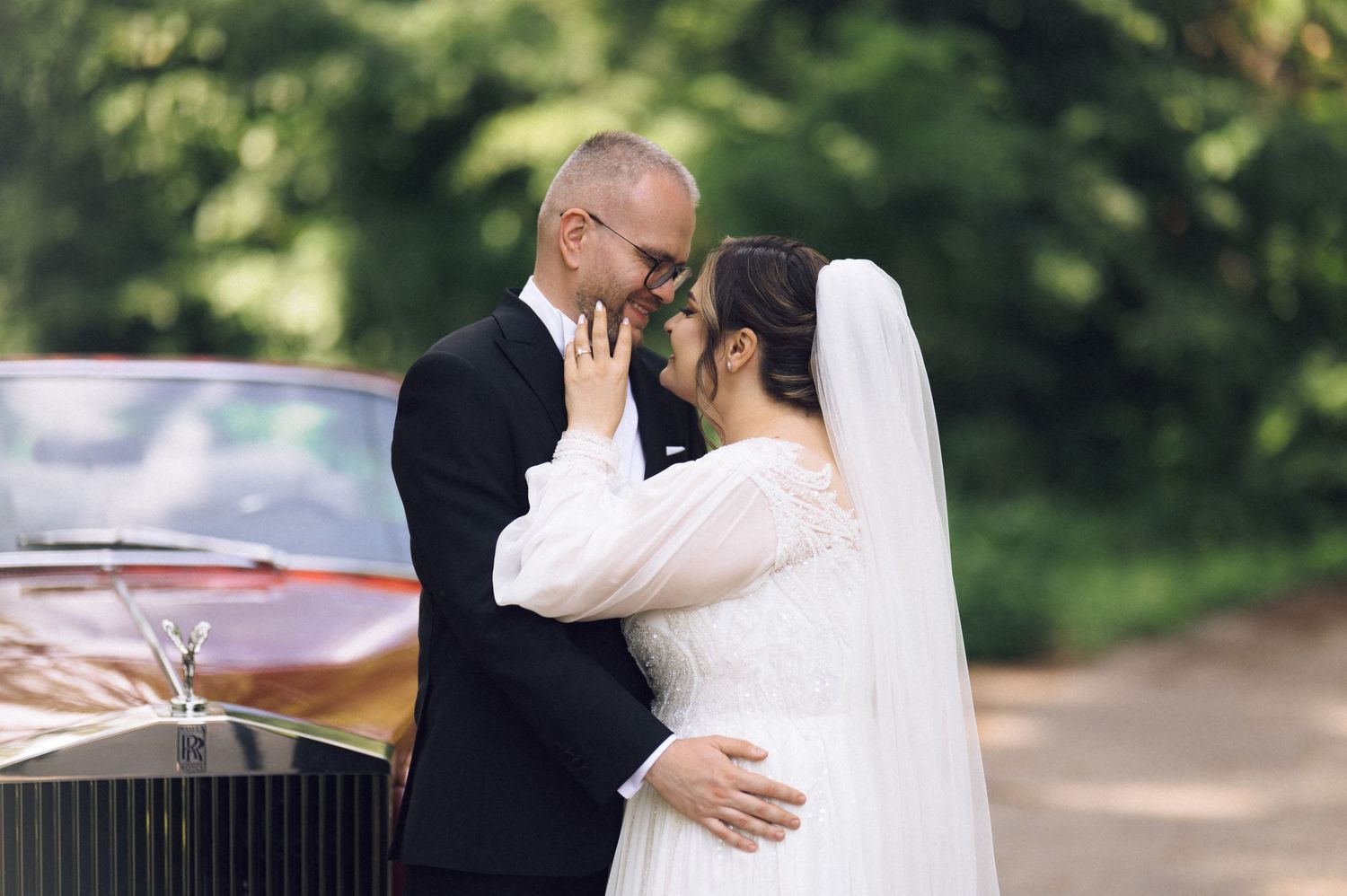 Newlywed couple sharing a romantic moment in front of a vintage car, bride in white gown and veil, groom in black suit.
