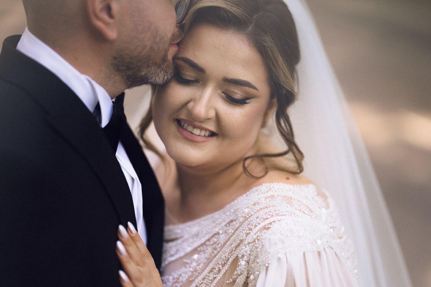A smiling bride in a beaded gown with veil receives a kiss from groom in black tuxedo on their wedding day.