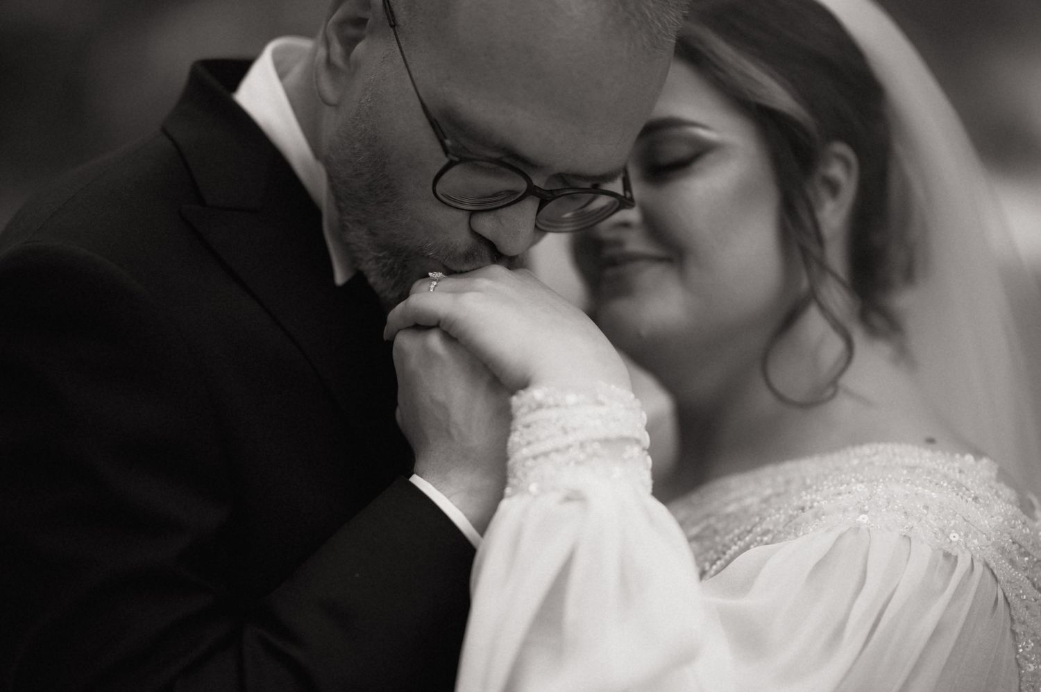 Black and white wedding photo of groom kissing bride's hand tenderly, both smiling with eyes closed.