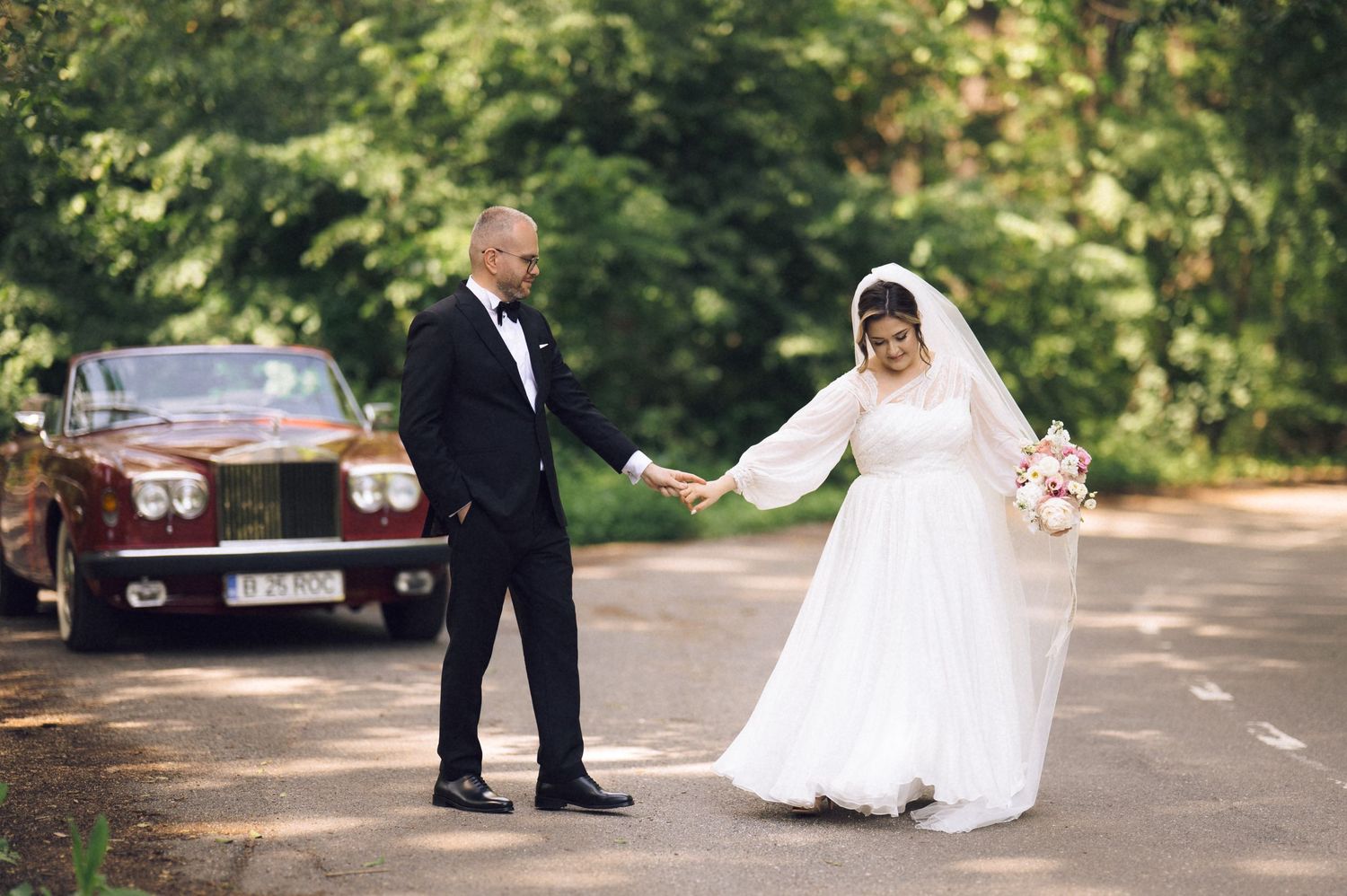 Bride and groom holding hands on a road, with a vintage red Rolls-Royce and lush green trees in the background.