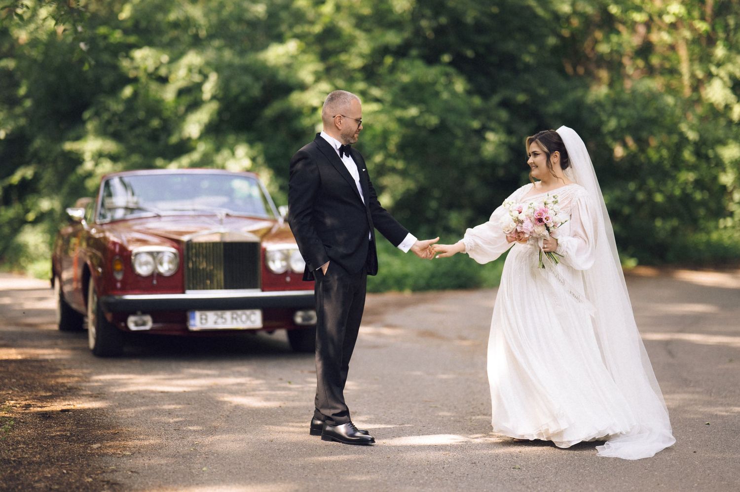 Bride and groom holding hands near a classic red Rolls-Royce on a tree-lined road during their wedding day.