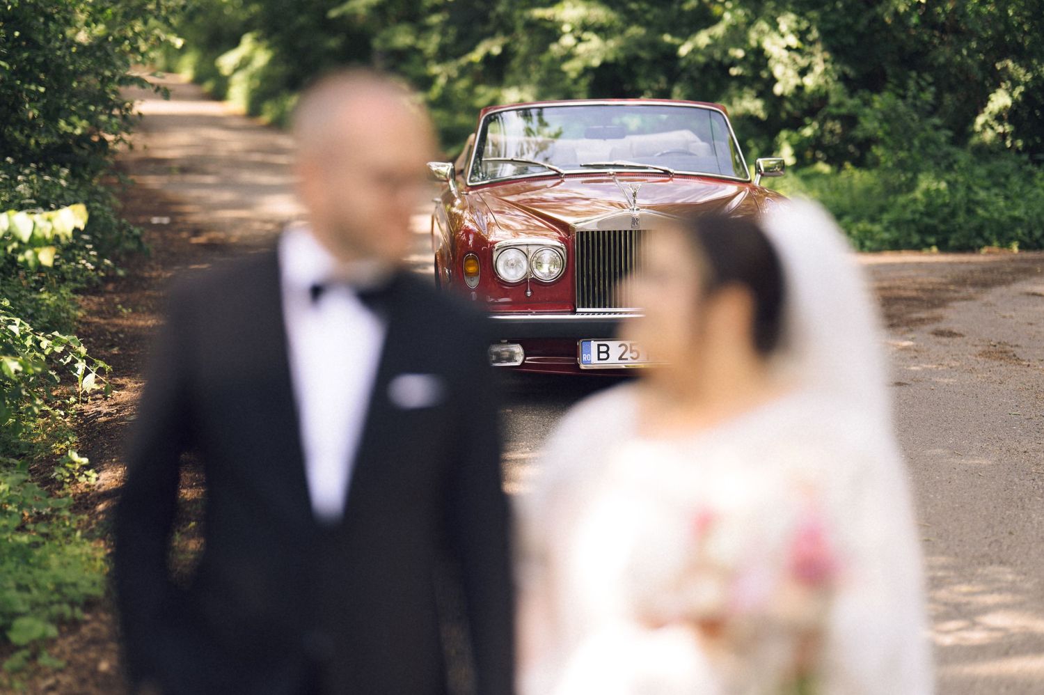 Bride and groom posing in front of a classic red Rolls-Royce on a tree-lined road at a wedding.