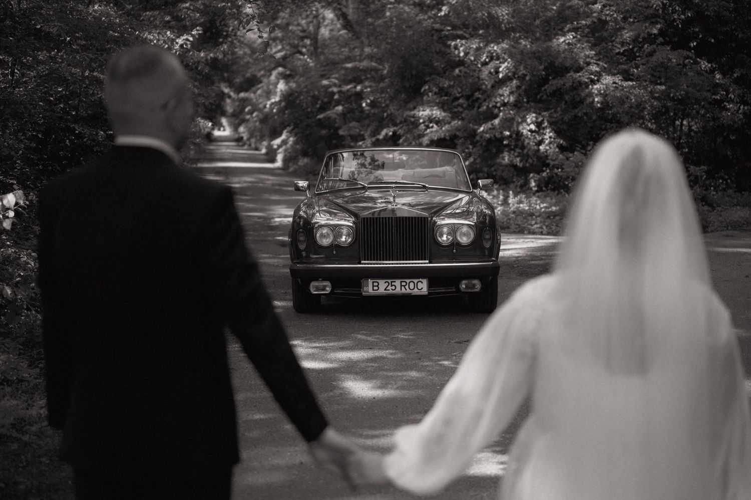 Black and white photo of bride and groom holding hands as a classic Rolls-Royce approaches on a tree-lined road.