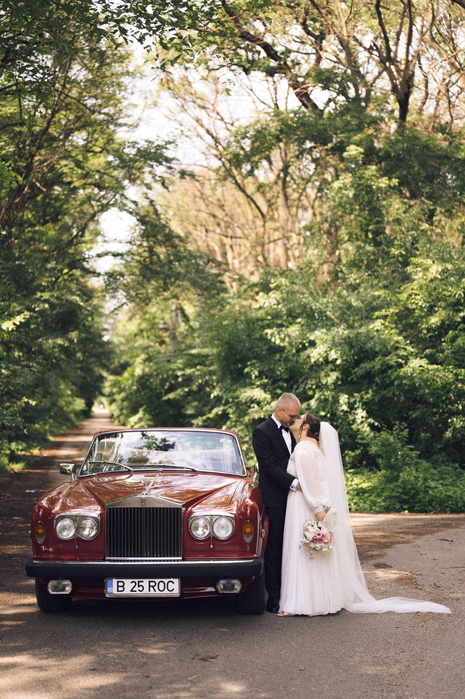 Bride and groom kissing beside a classic red Rolls-Royce on a tree-lined road on their wedding day.