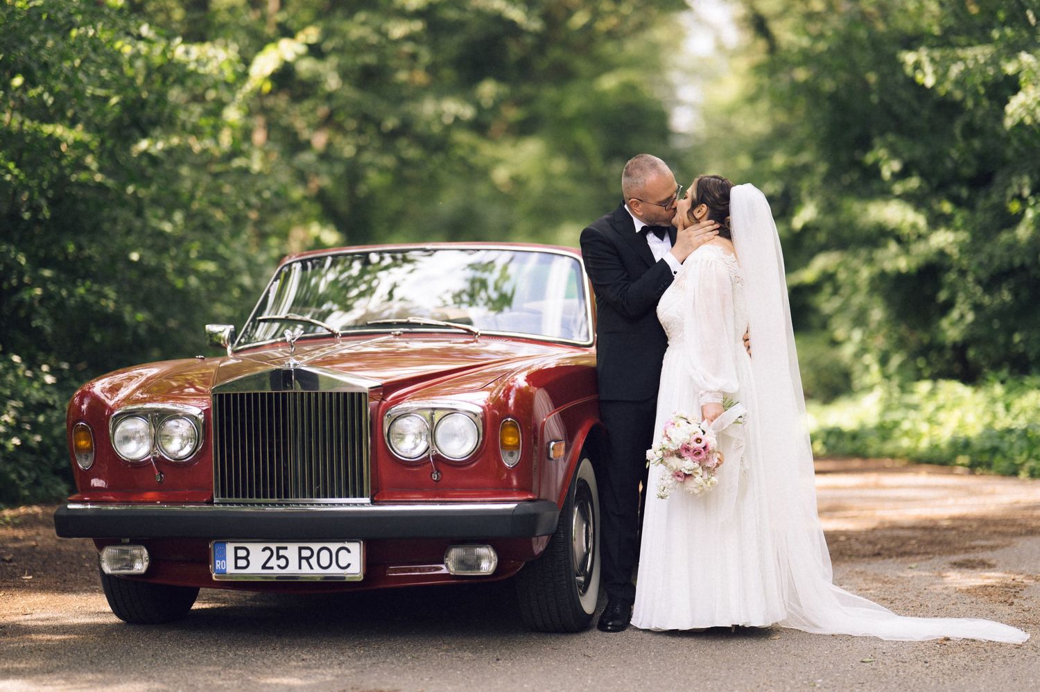 Bride and groom kissing beside a classic red Rolls-Royce on a tree-lined road on their wedding day.