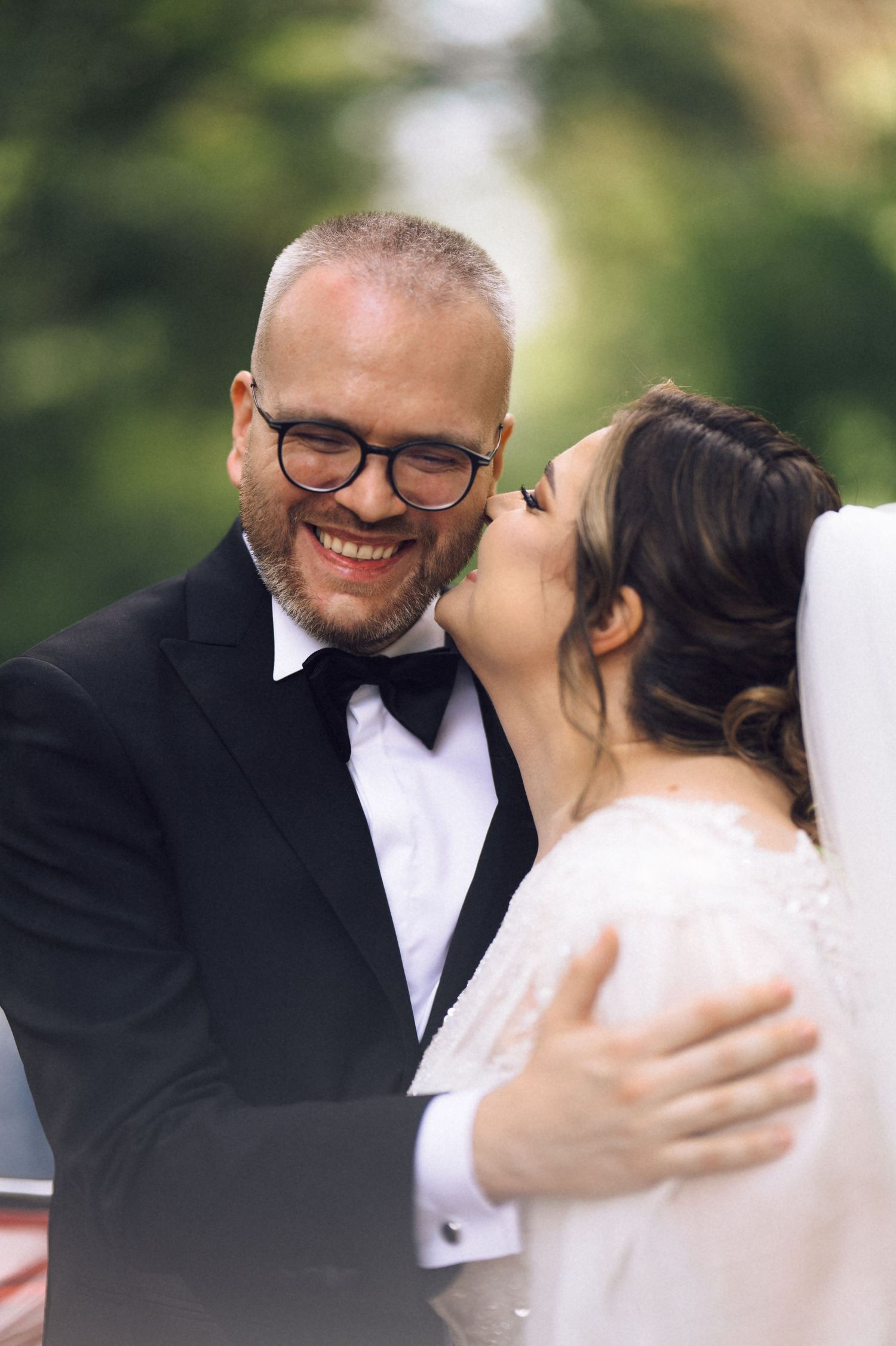 Smiling groom in black tuxedo and glasses receives a kiss on the cheek from bride in white wedding dress outdoors.