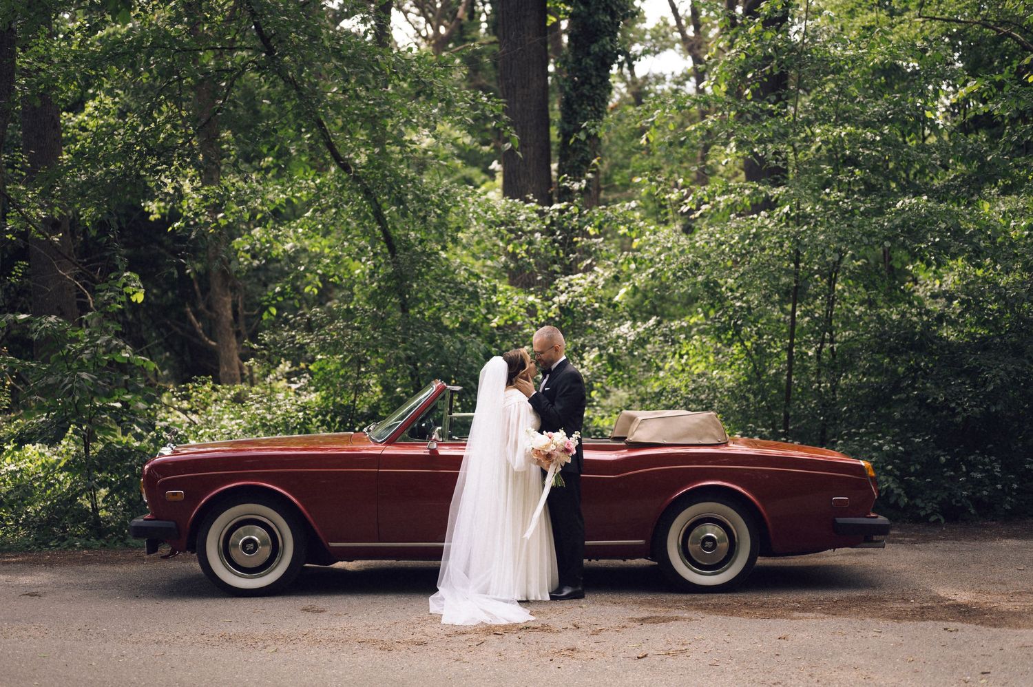 Bride and groom kissing beside a classic red convertible car surrounded by lush green trees on their wedding day.