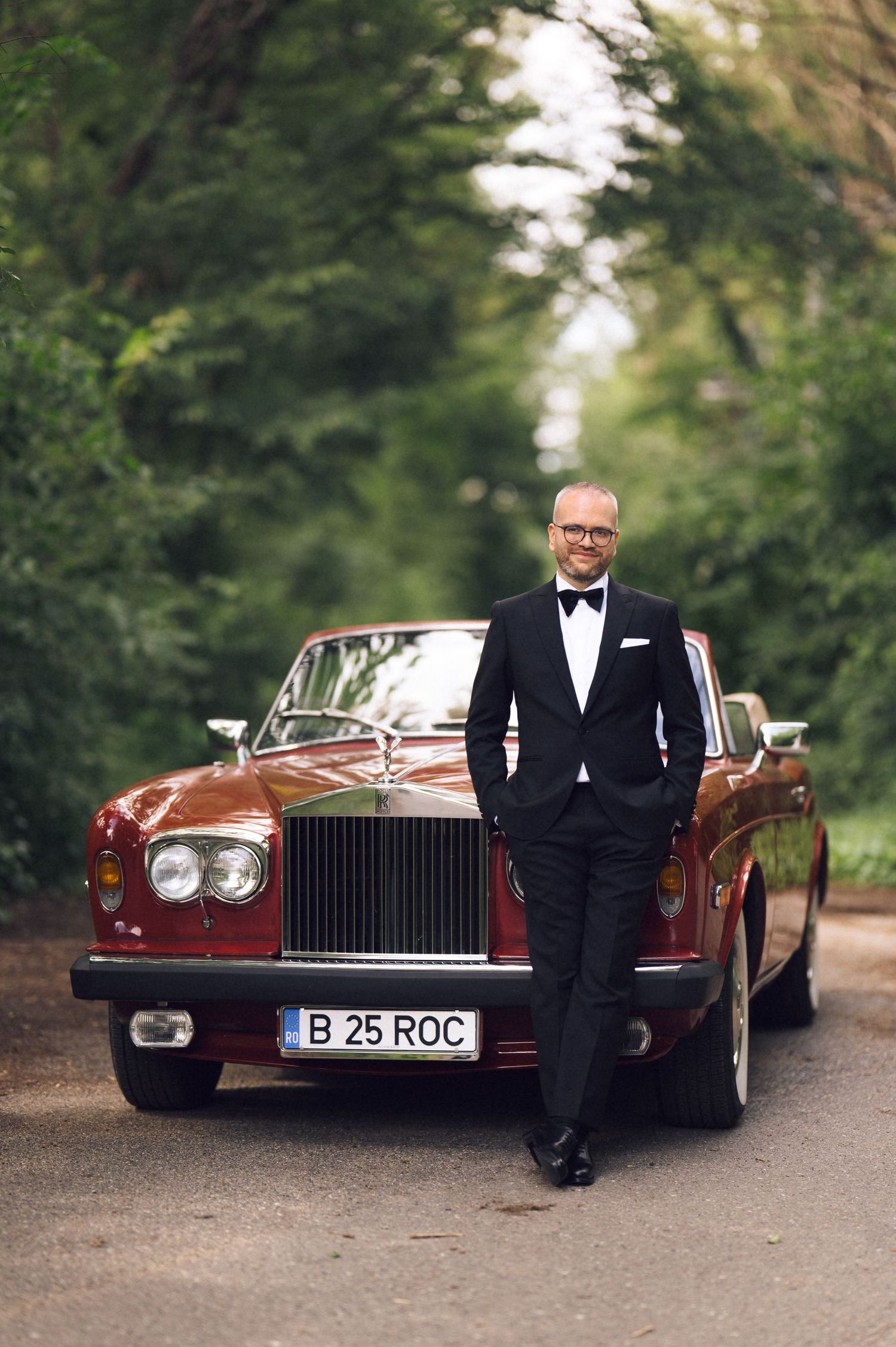 Man in tuxedo posing beside a classic burgundy Rolls-Royce with Romanian license plate on a tree-lined road.