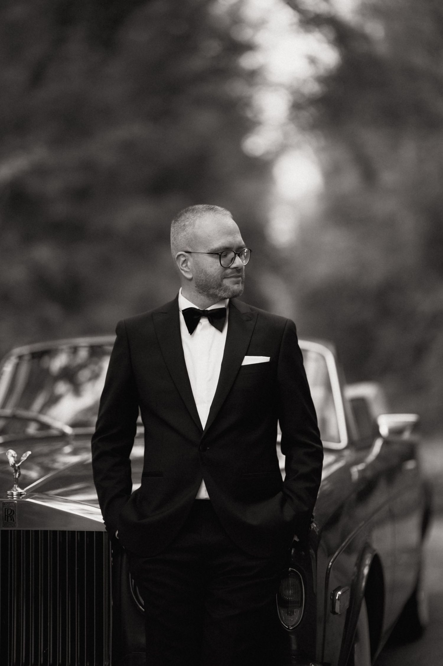 A man in a tuxedo with bow tie and glasses leans against a vintage car in a black and white portrait.