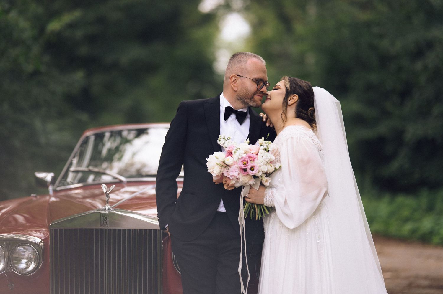 Bride and groom share a romantic moment beside a vintage red Rolls-Royce, surrounded by lush green trees.