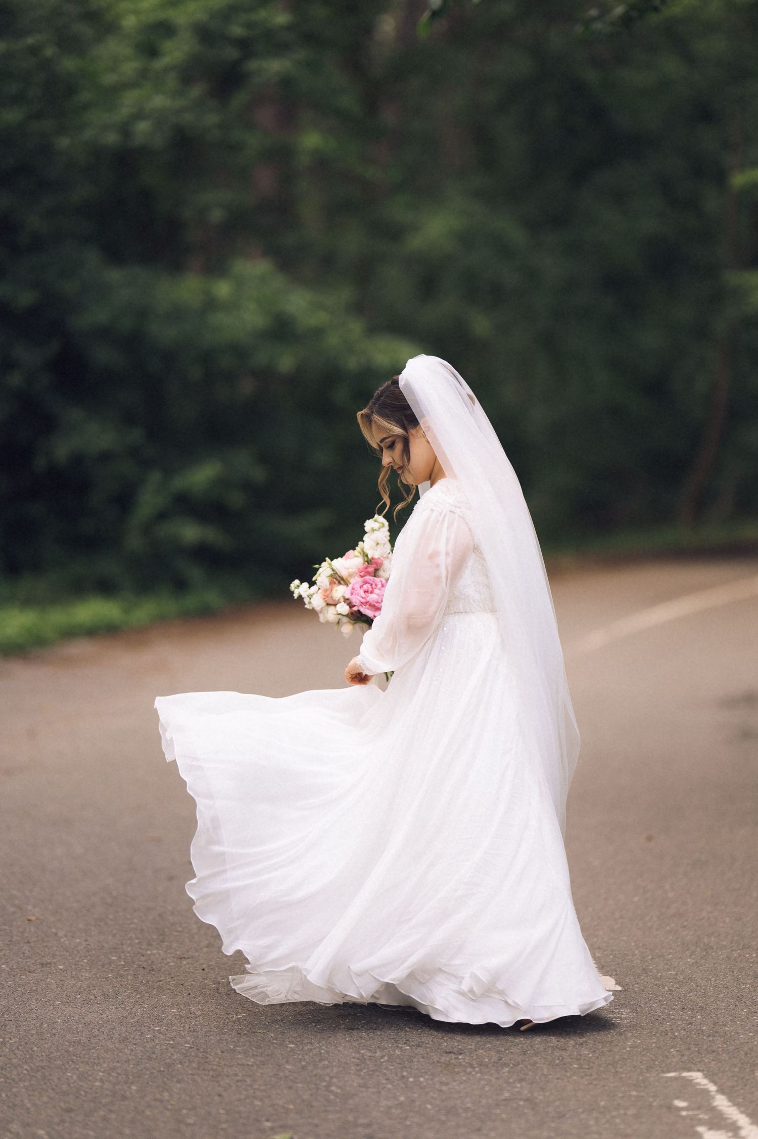 Bride in white gown and veil twirls on road holding pink bouquet with lush green trees in background.