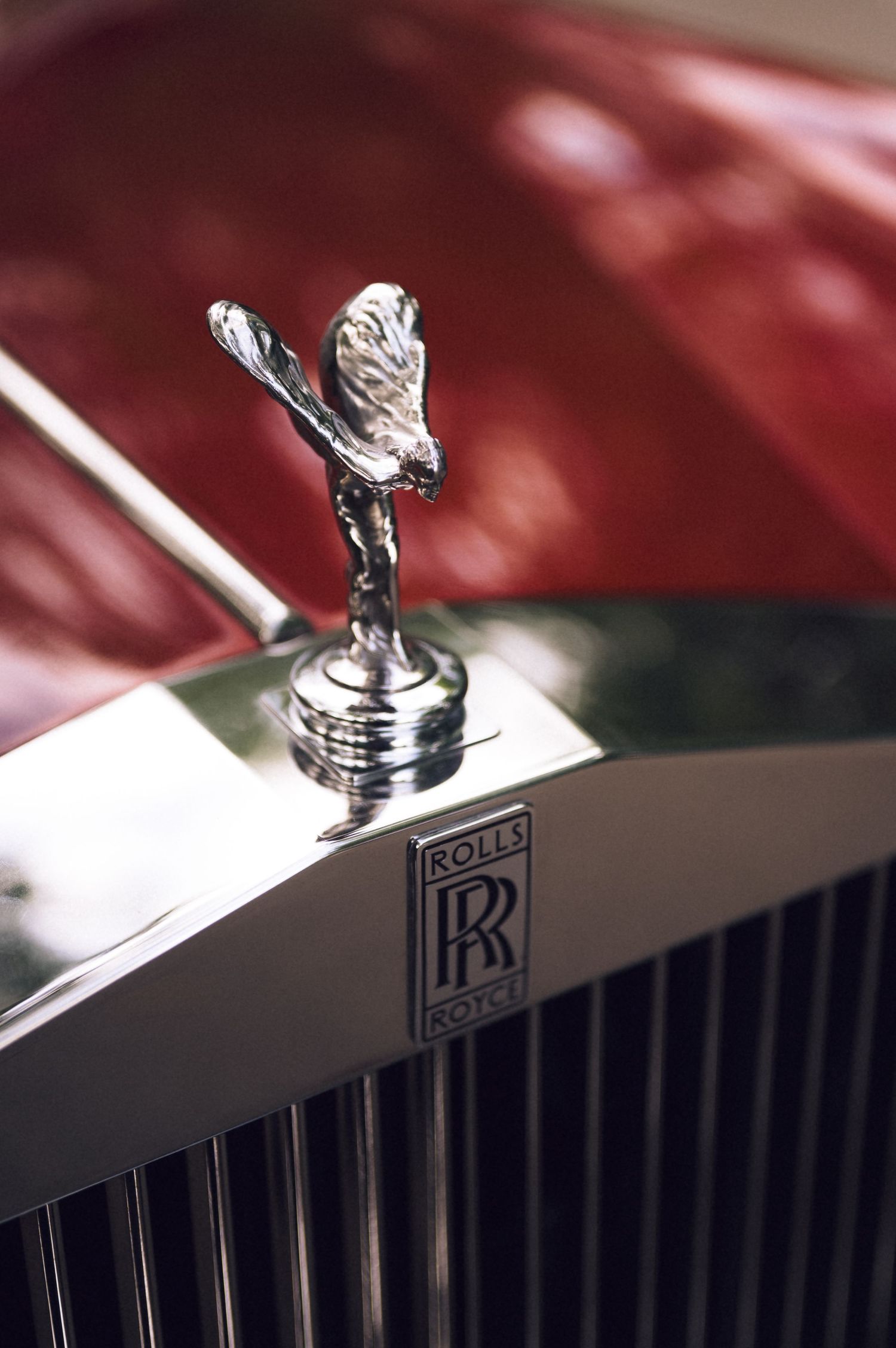 Close-up of a chrome Rolls-Royce Spirit of Ecstasy hood ornament and RR logo on a red luxury car.