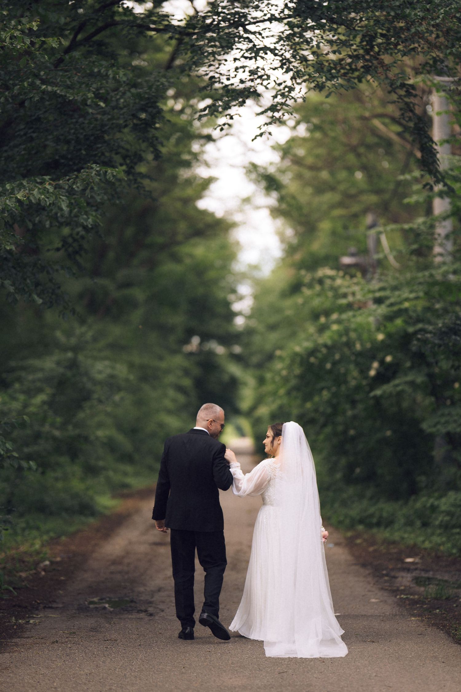 A bride and groom walk hand in hand down a tree-lined path, the bride in a white gown with veil.