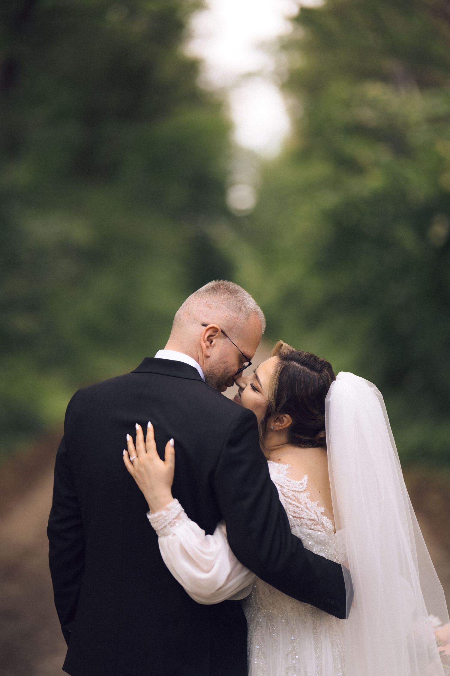 A bride and groom share an intimate moment on a tree-lined path, bride in lace gown and veil, groom in black suit.