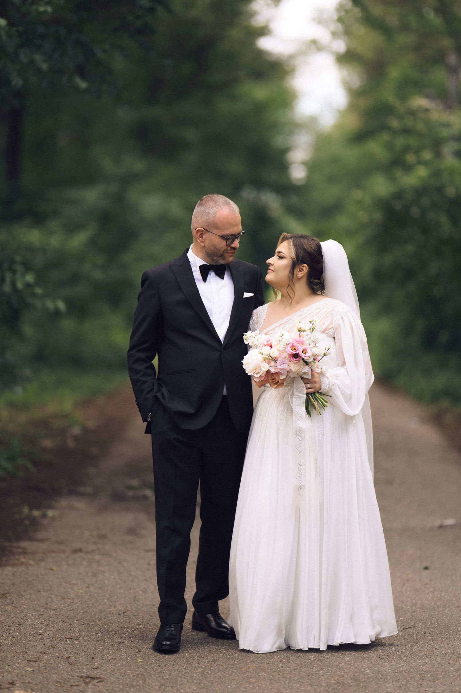 A bride in a white gown and groom in a black tuxedo walk together on a tree-lined path on their wedding day.