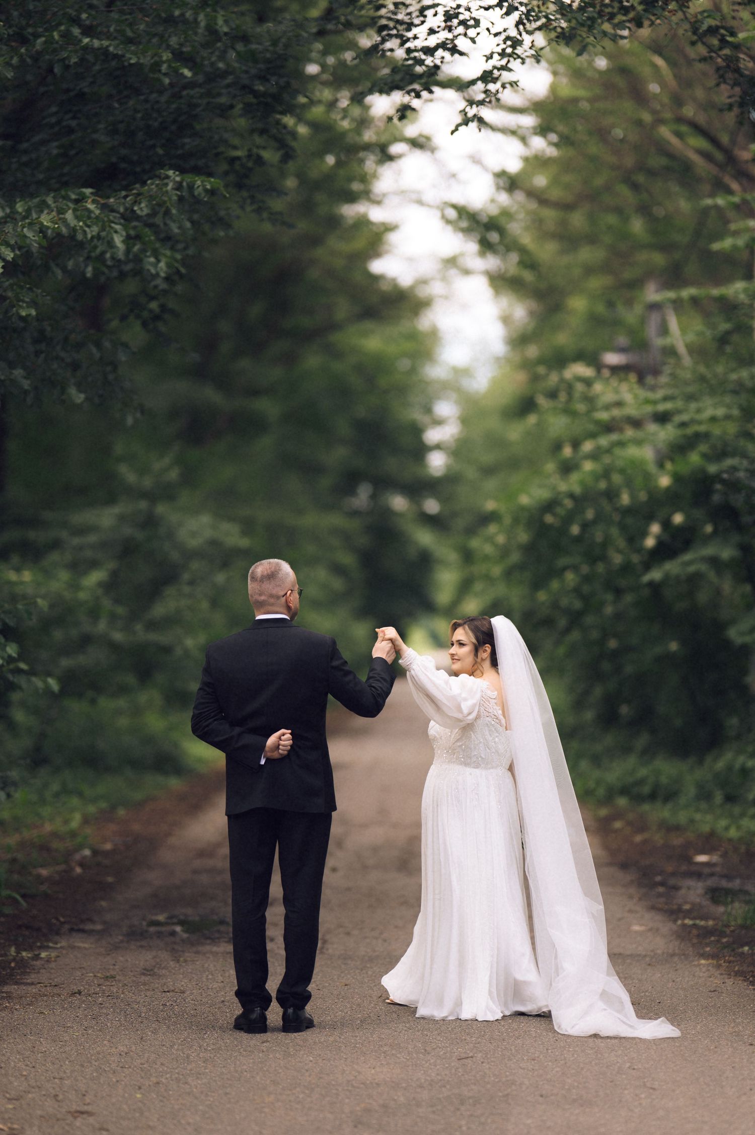 Bride and groom holding hands on a tree-lined path, bride in white gown with veil, groom in black tuxedo.