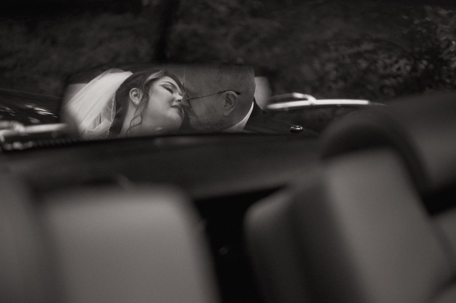 Black and white photo of a bride and groom sharing a romantic kiss inside a car on their wedding day.