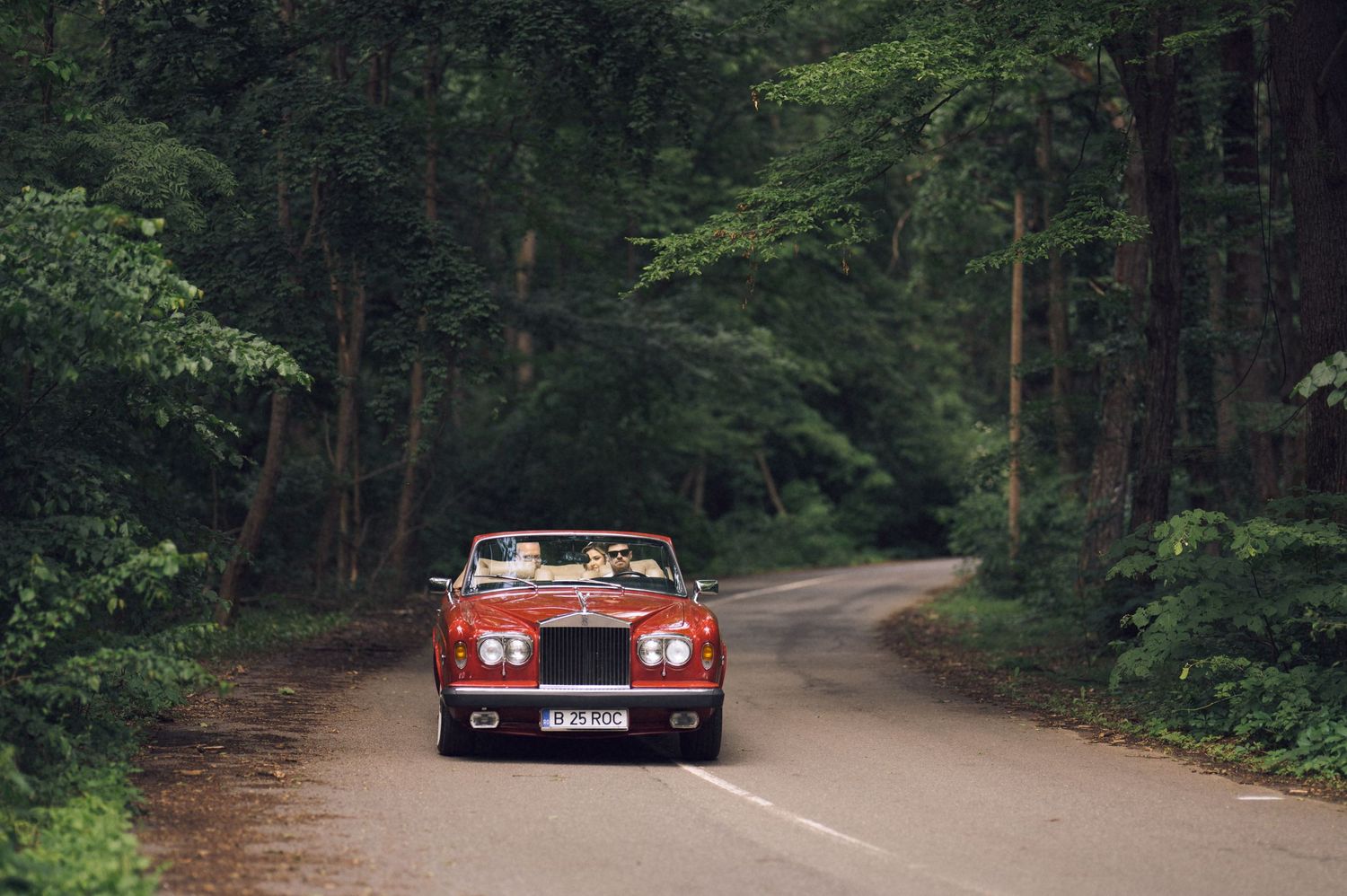 Red convertible Bentley driving on a narrow road surrounded by lush green forest trees.