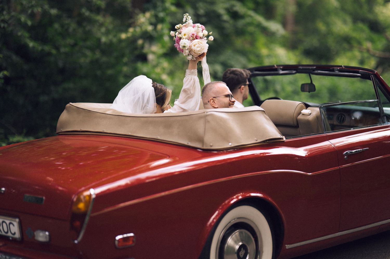 Bride holding bouquet aloft in red Rolls-Royce convertible with groom, wedding car photo.