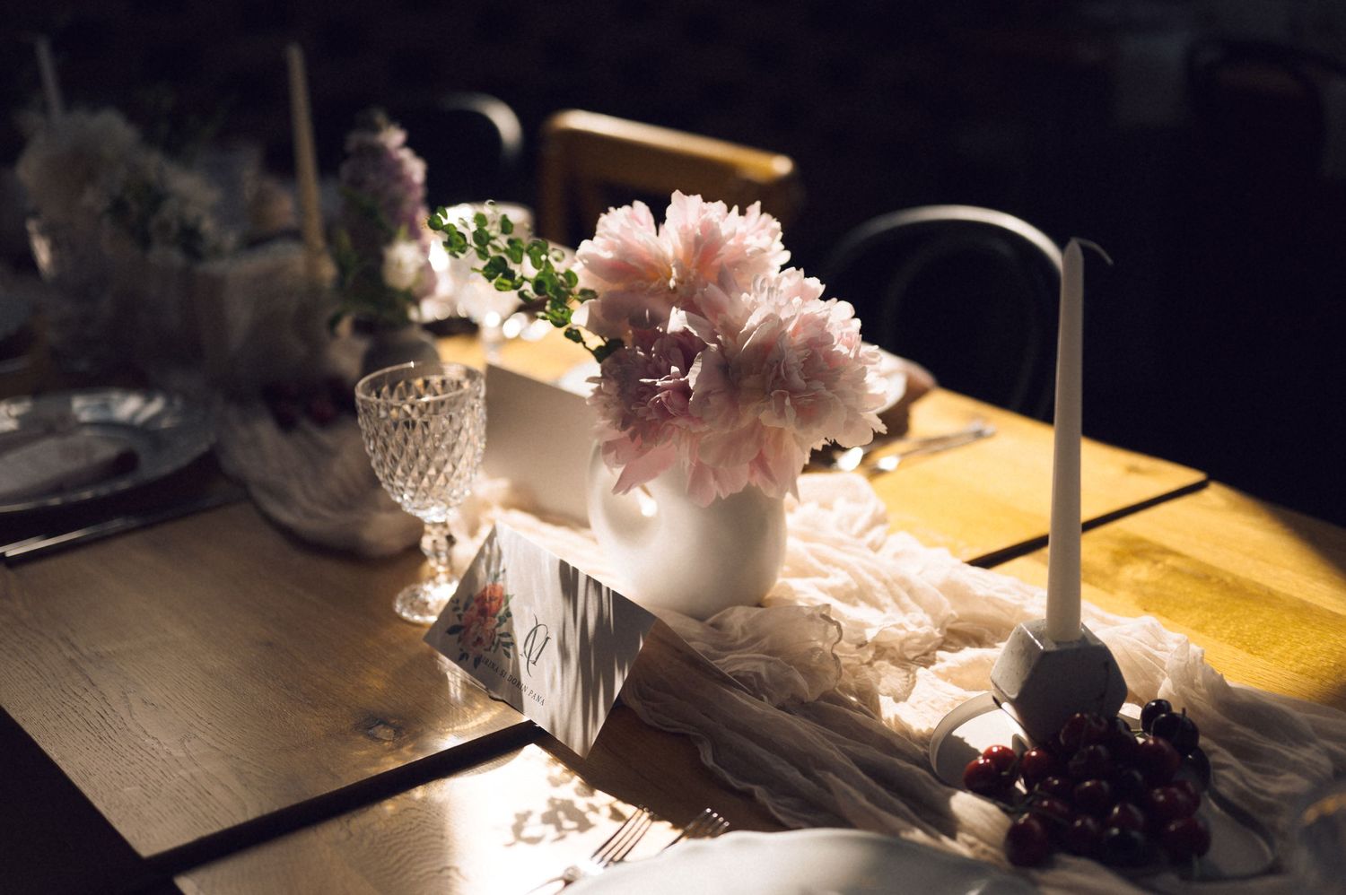 Elegant wedding table setting with pink peonies in white vase, crystal glassware, and candles on rustic wooden table.