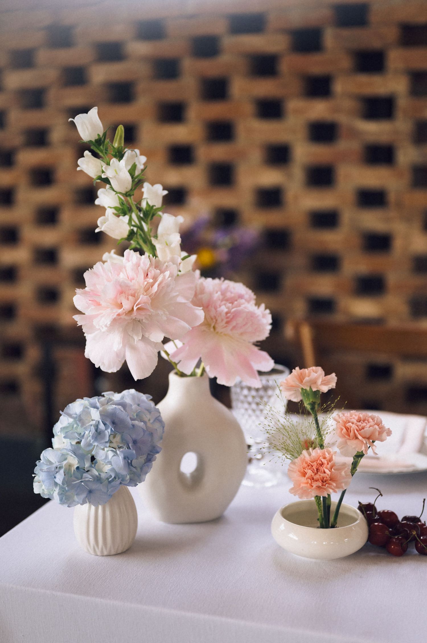 Three white ceramic vases with pink carnations, blue hydrangeas, and blush blooms arranged on a white table.