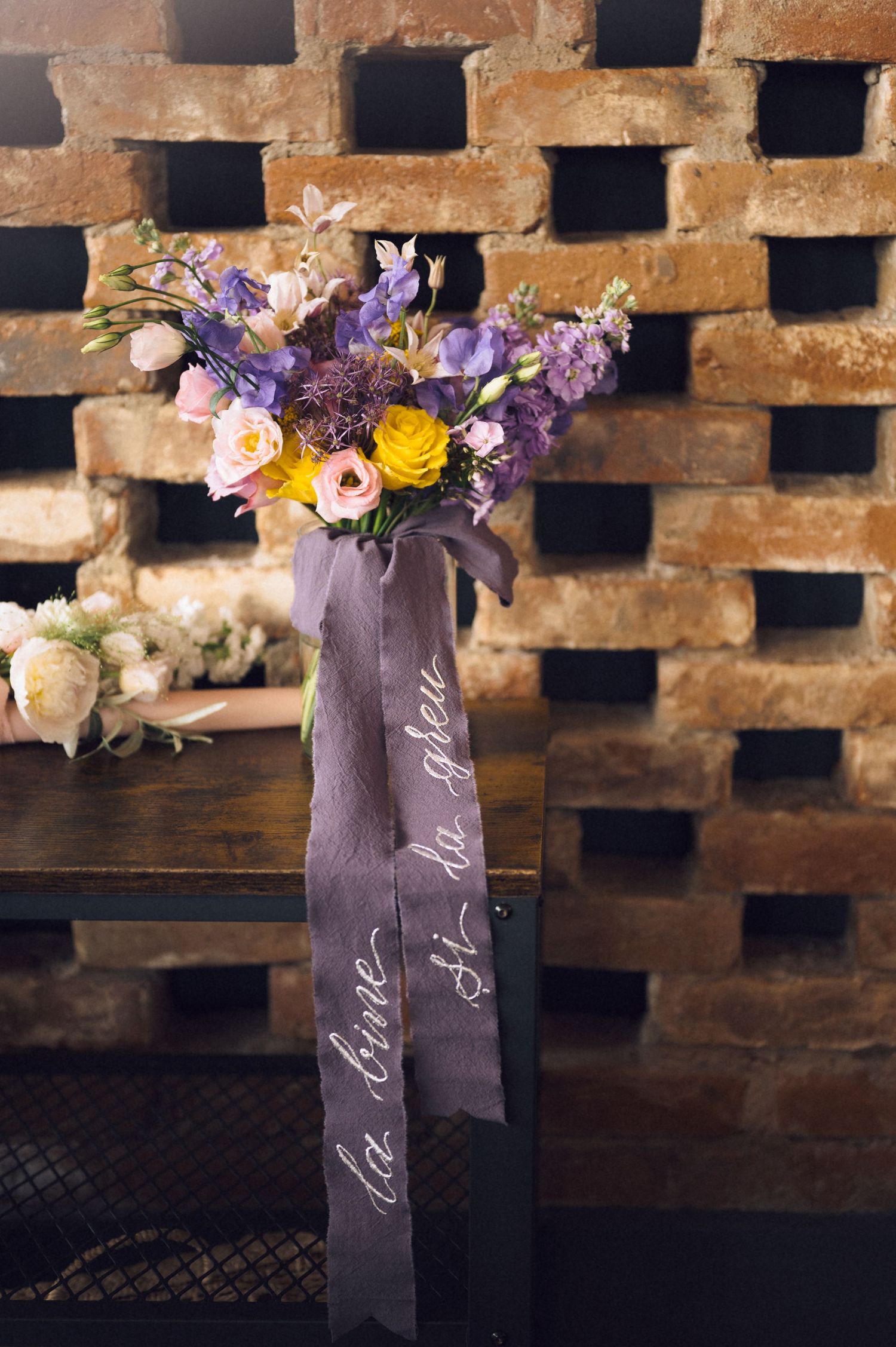 Purple and yellow floral bouquet with calligraphy ribbon on dark wood bench against brick wall.