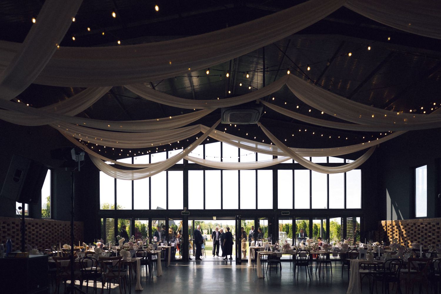 Elegant wedding reception hall with draped white fabric ceiling, string lights, and large windows overlooking lush greenery.