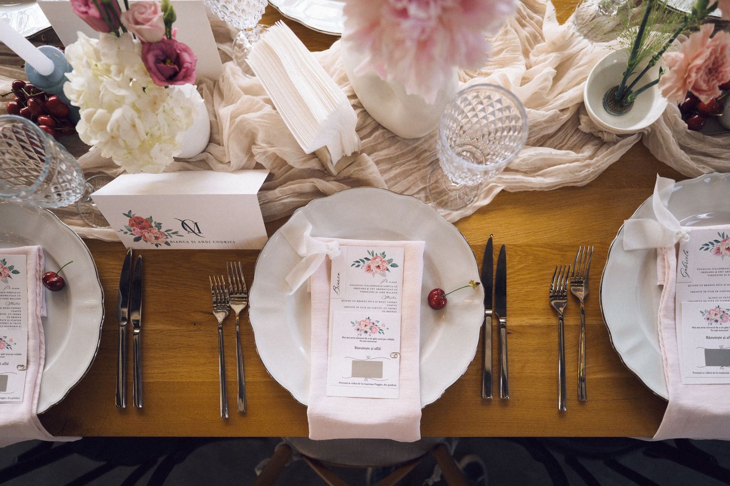Elegant wedding table setting with floral arrangements, white plates, silverware, and pink menu cards on a wooden table.