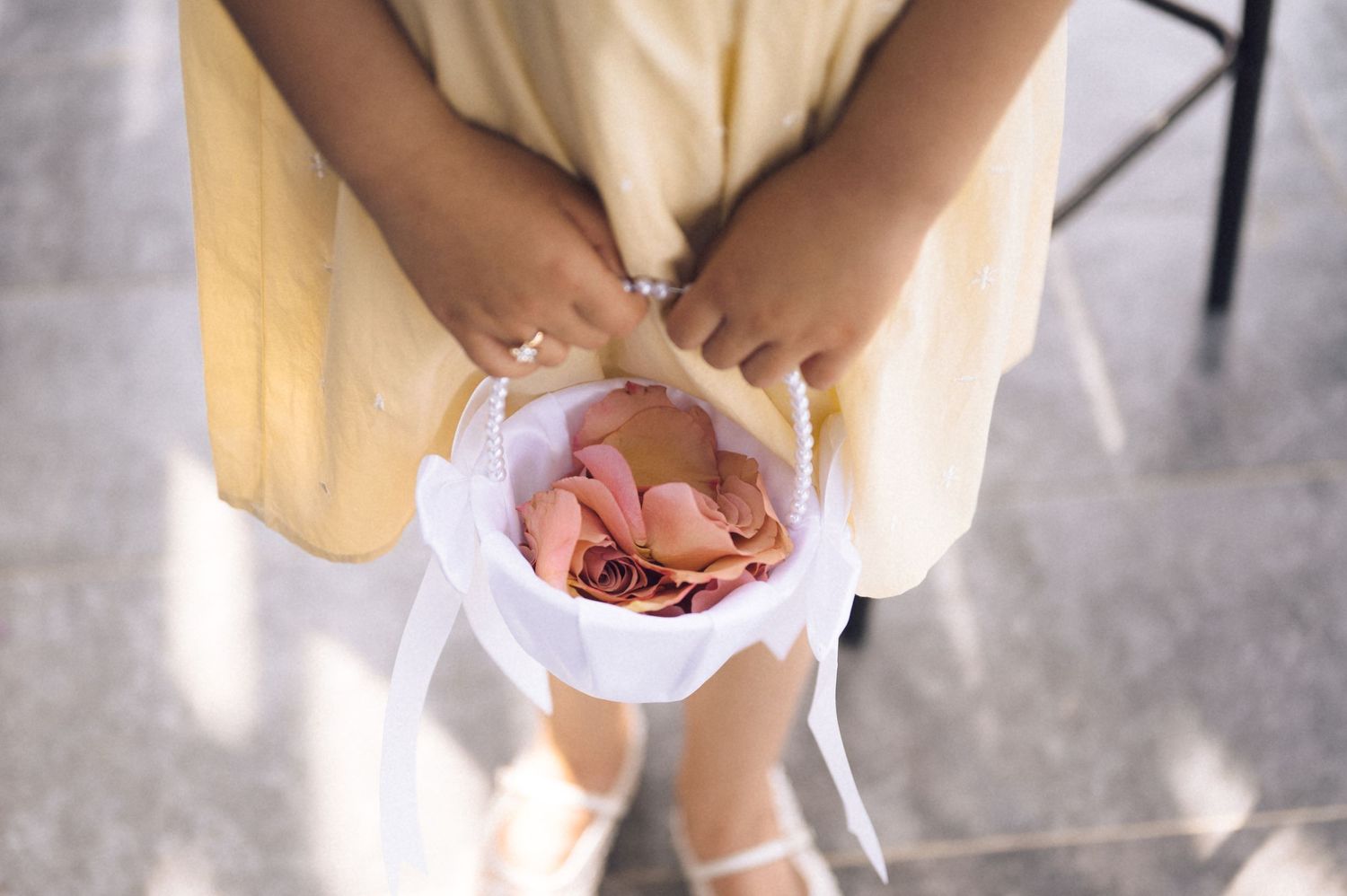 Flower girl in yellow dress holding white basket filled with pink rose petals at a wedding ceremony.