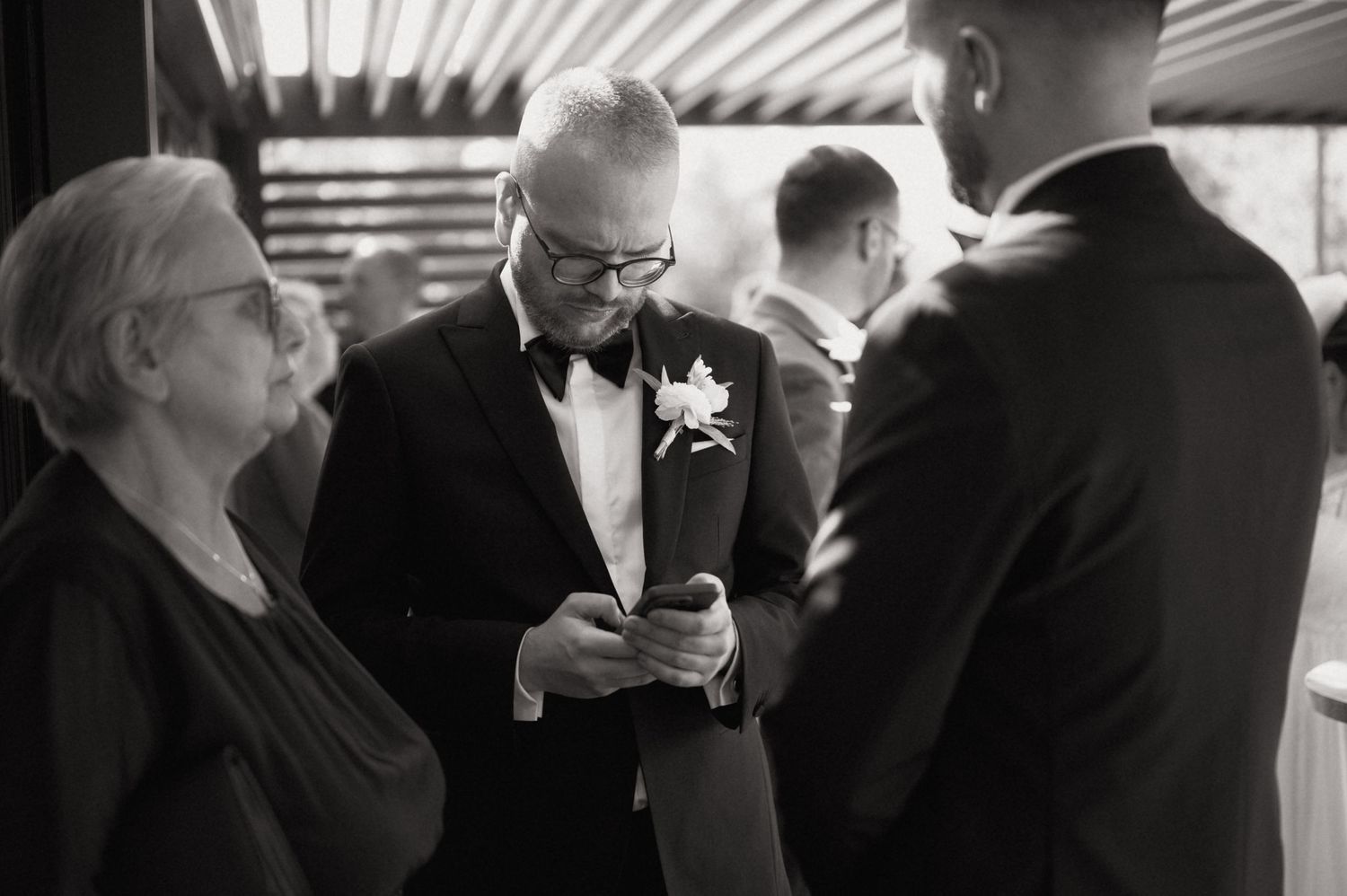 Black and white wedding photo of elegantly dressed guests in tuxedos examining something at a formal reception.