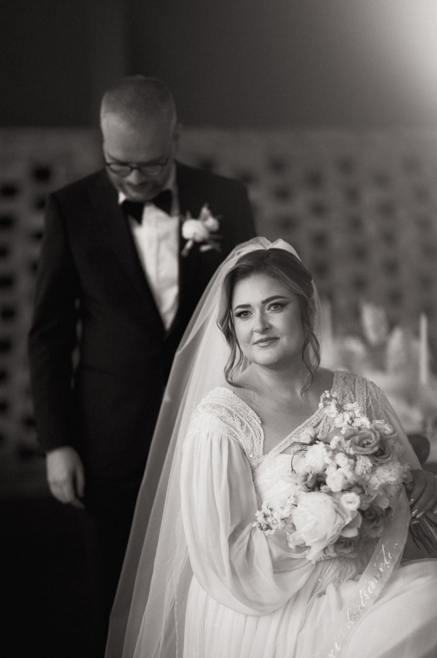 Black and white wedding photo of a bride in a lace gown holding a bouquet, standing before a man in a tuxedo.