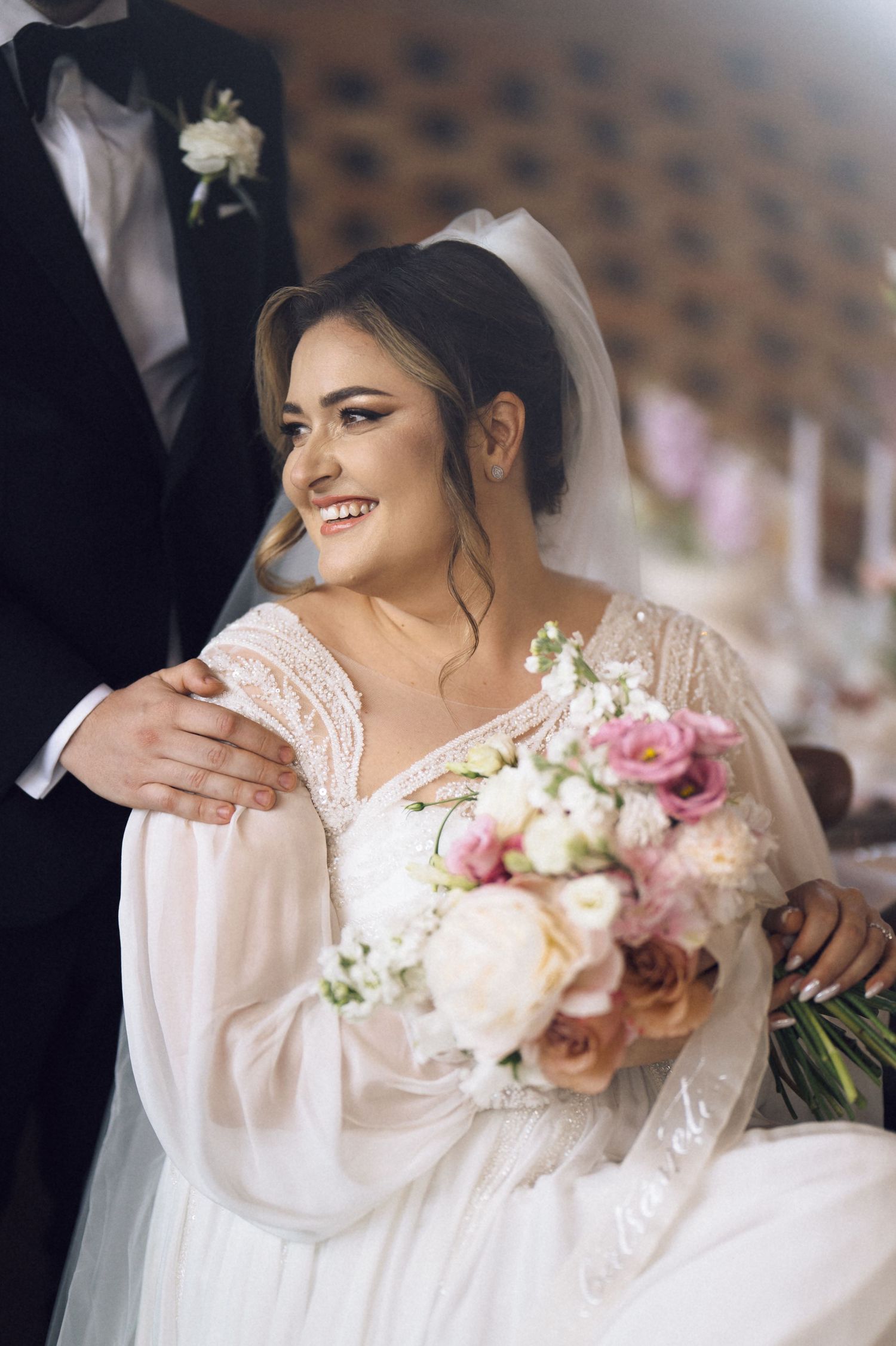 Beautiful smiling bride in white lace gown holding pink and white bouquet while groom places hand on her shoulder.