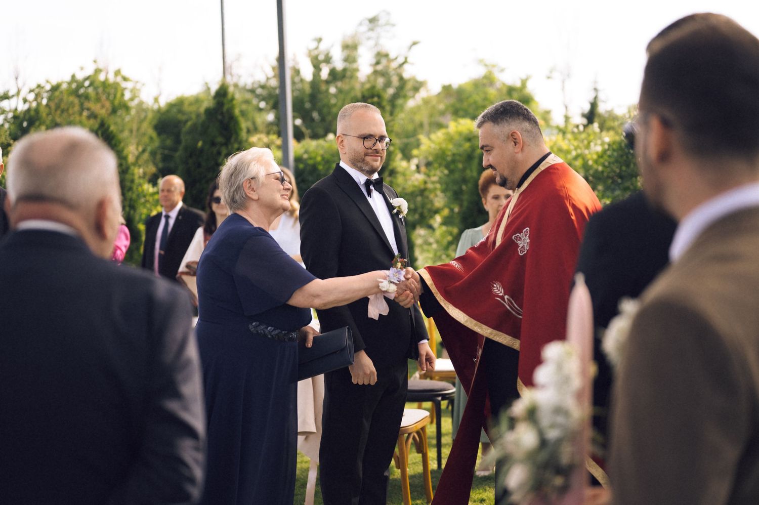 Outdoor wedding ceremony with guests shaking hands with a priest in red vestments on a sunny day.