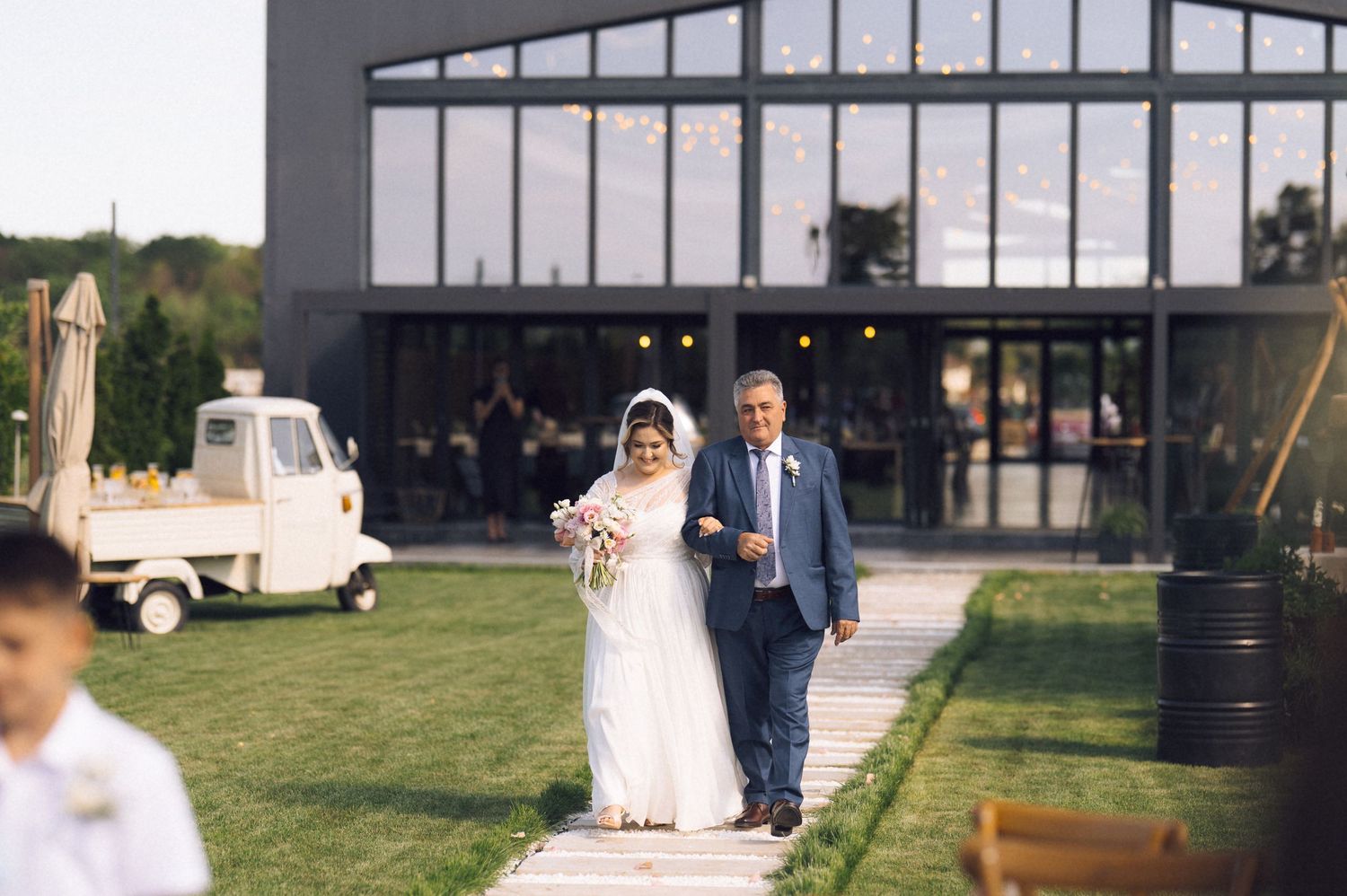 Bride and groom walking down outdoor wedding aisle toward modern venue with string lights at elegant reception.